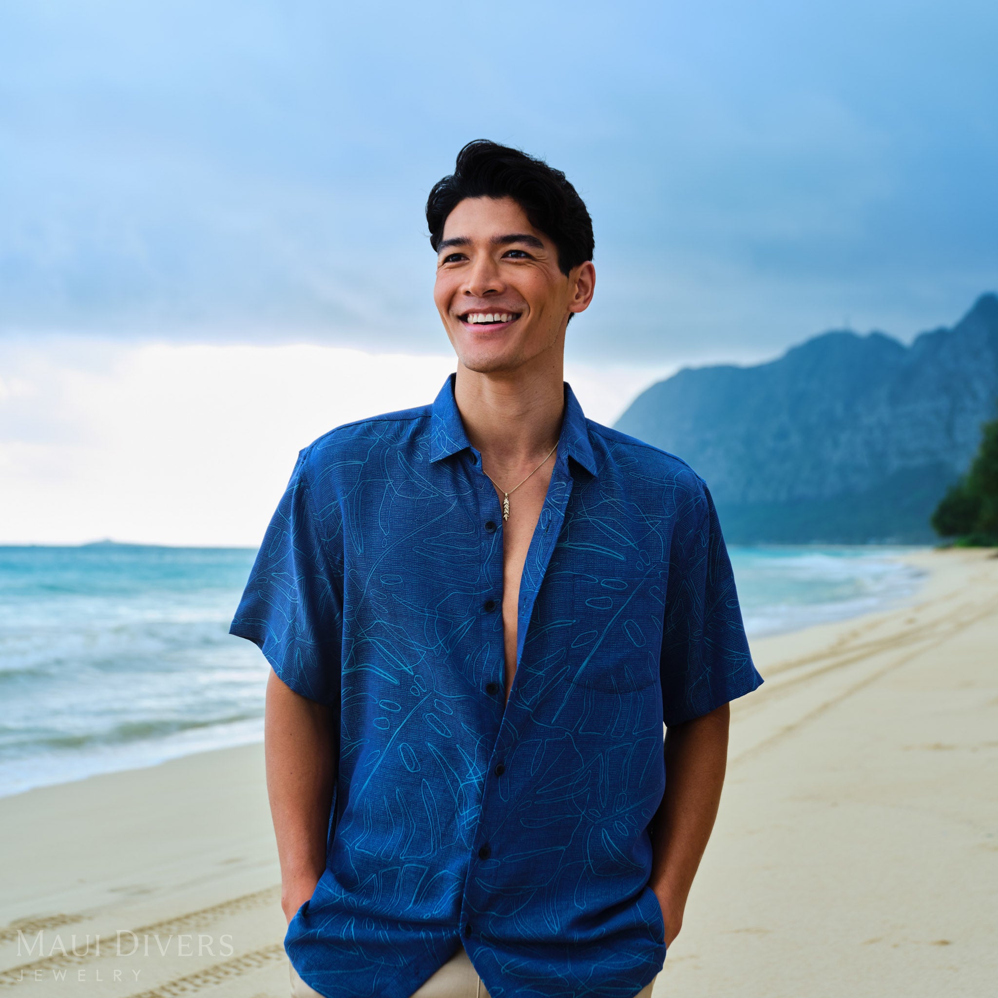 Man wearing a blue patterned shirt with a 35mm Maile Pendant in Gold on a beach with mountains in the background