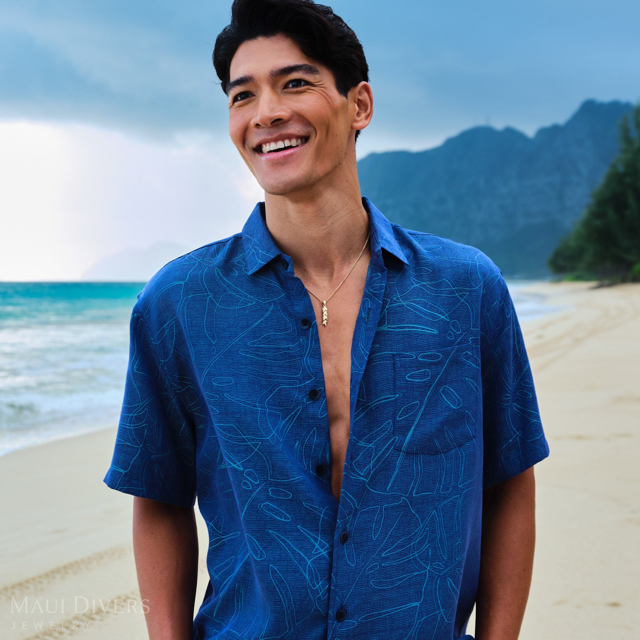Man wearing a blue patterned shirt with a 35mm Maile Pendant in Gold on a beach with mountains in the background