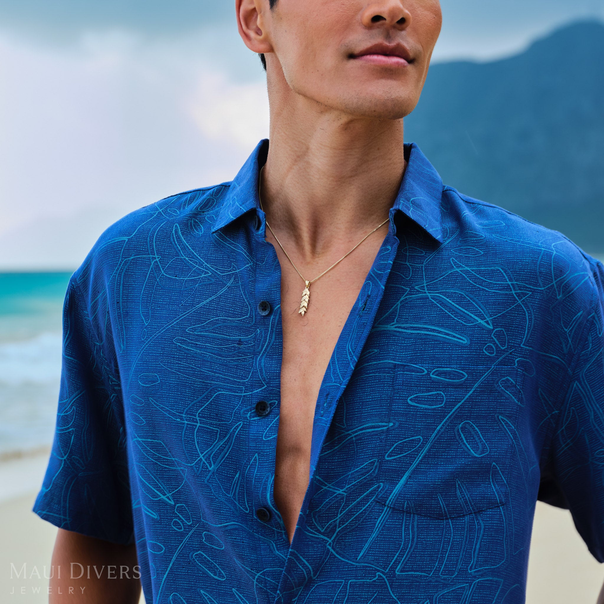 Man wearing a blue patterned shirt with a gold maile leaf pendant on a chain on a beach with mountains in the background