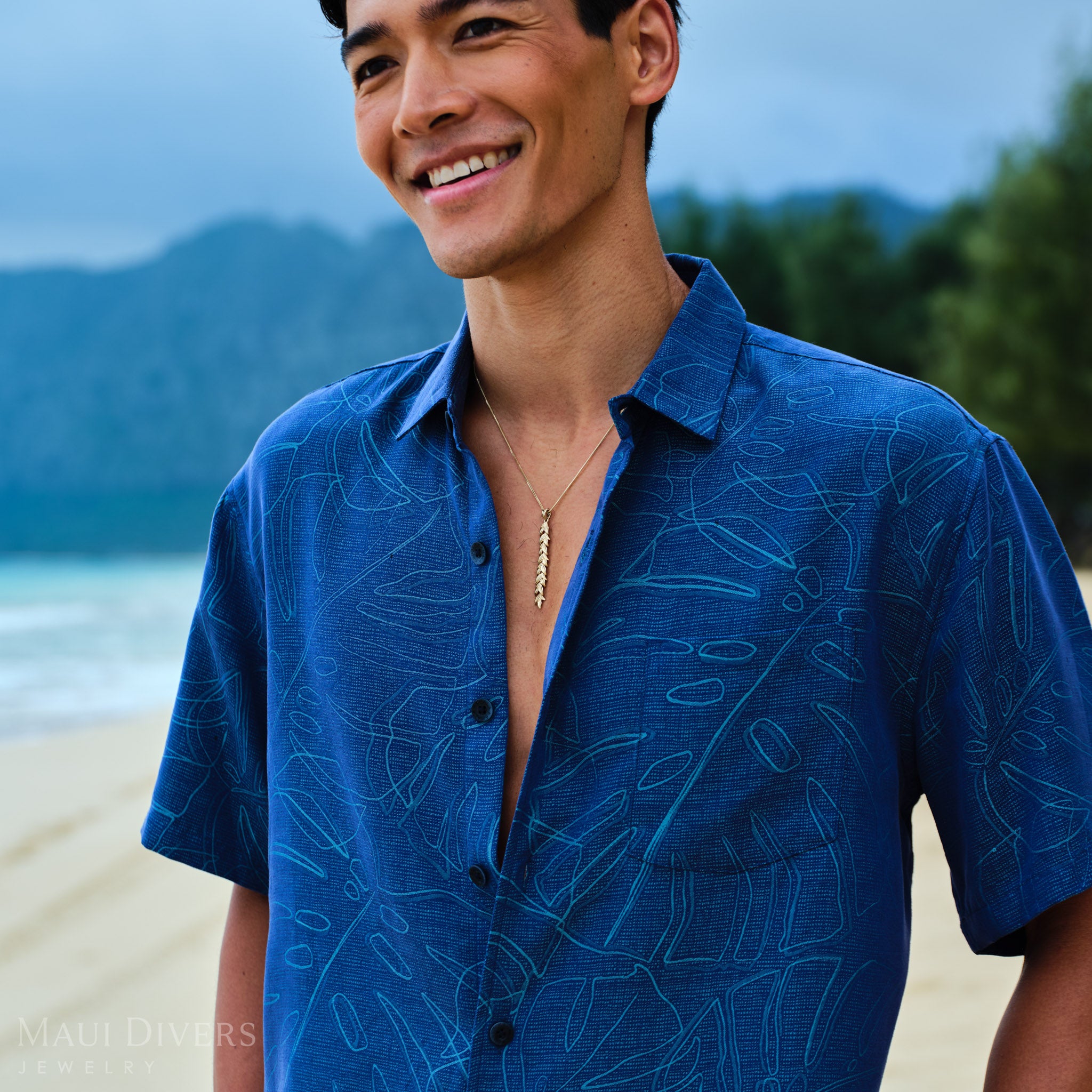 Man wearing a 58mm Maile Pendant in Gold and blue patterned shirt on a beach with mountains in the background