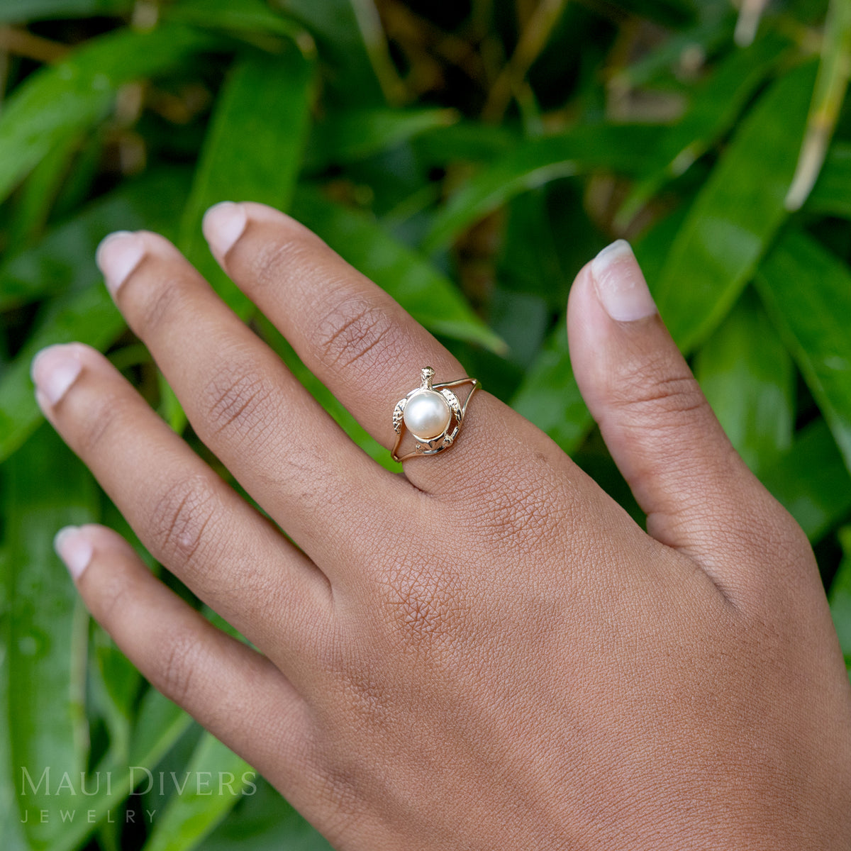 A hand wearing a Pick A Pearl Honu Ring in 14k yellow gold with a freshwater white pearl, against a blurred leafy green background