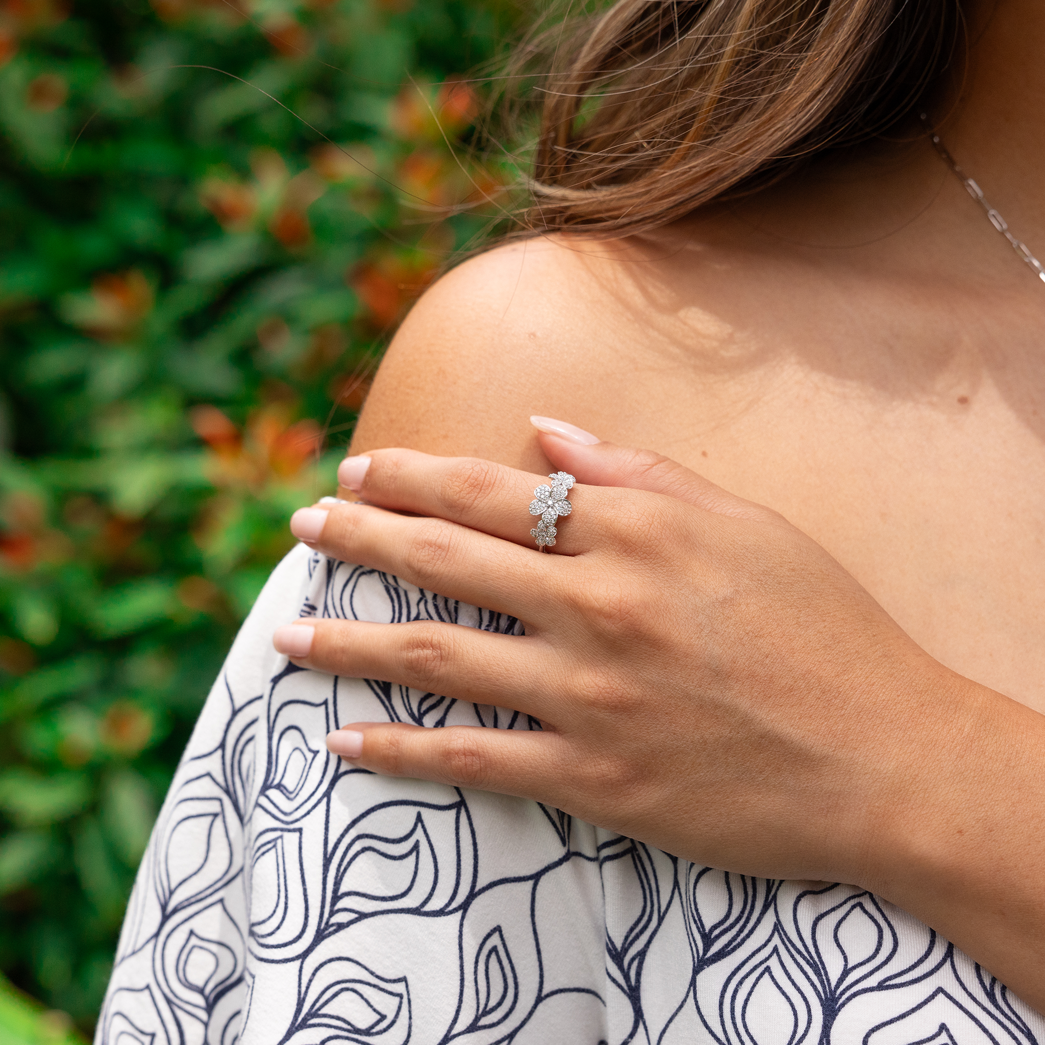 Close up of a 10mm Three Plumeria Ring in White Gold with Diamonds on a hand with blurred greenery in the back