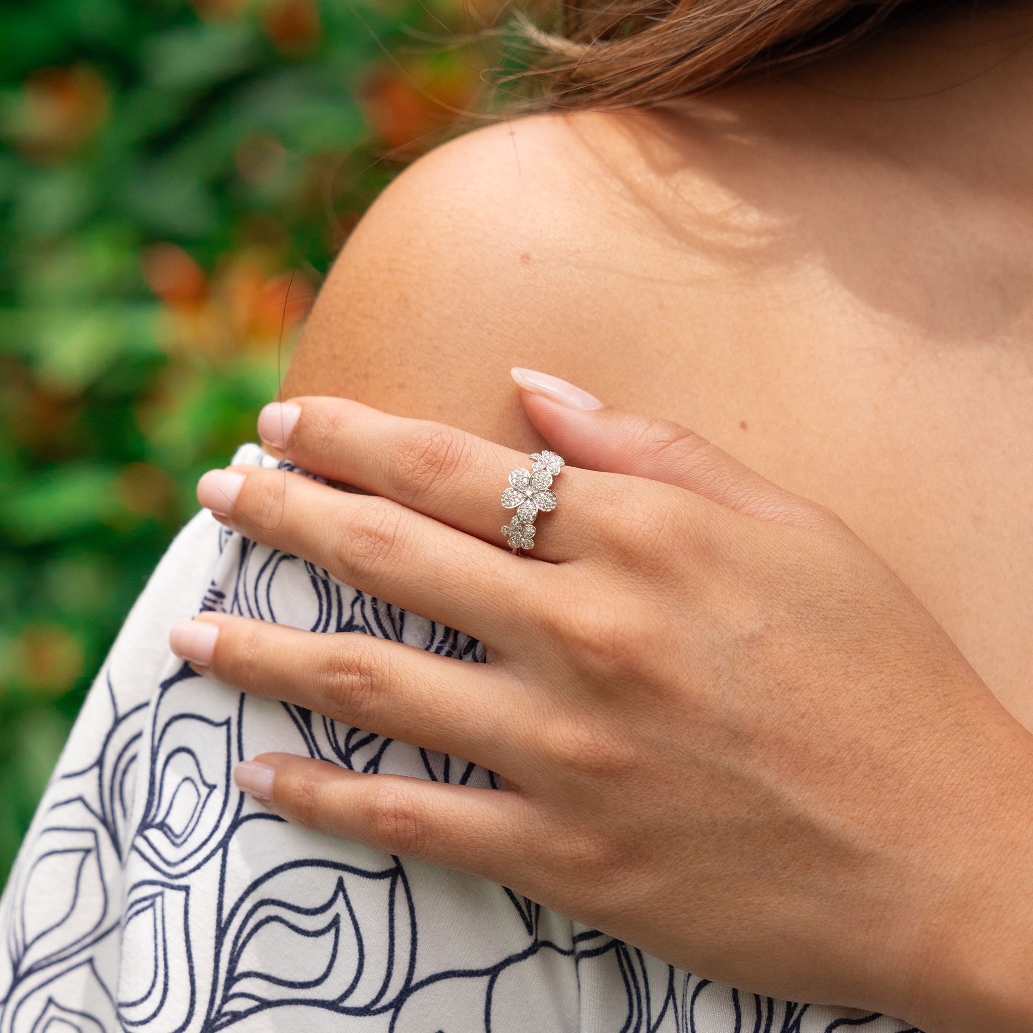 Close up of a 10mm Three Plumeria Ring in White Gold with Diamonds on a hand with blurred greenery in the back