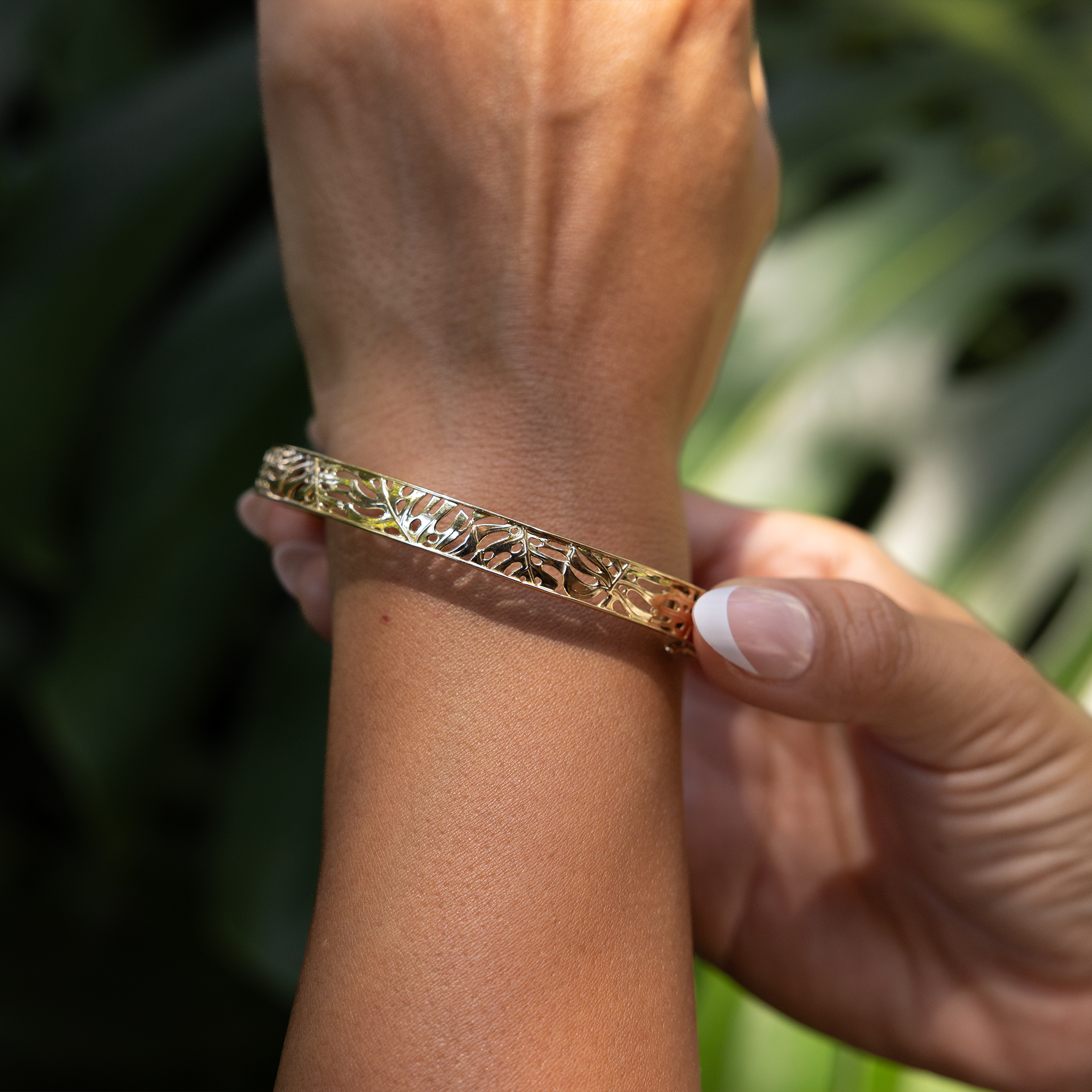 Close up of 8mm Monstera Hinge Bracelet in Gold on wrist with blurred monstera leaves in the background