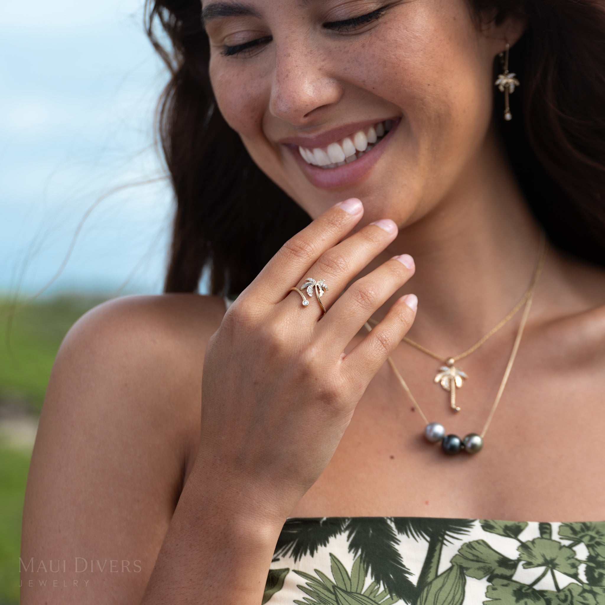 Close-up of a smiling woman wearing a gold and diamond palm tree bypass ring with a blurry sky and green plants in the background