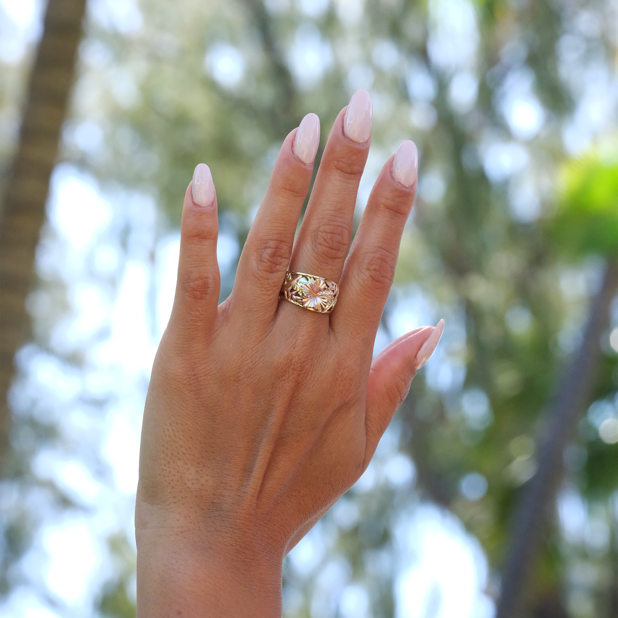 A woman's hand with a Hawaiian Gardens Hibiscus Ring in Tri Color Gold with Diamonds, blurred trees in background