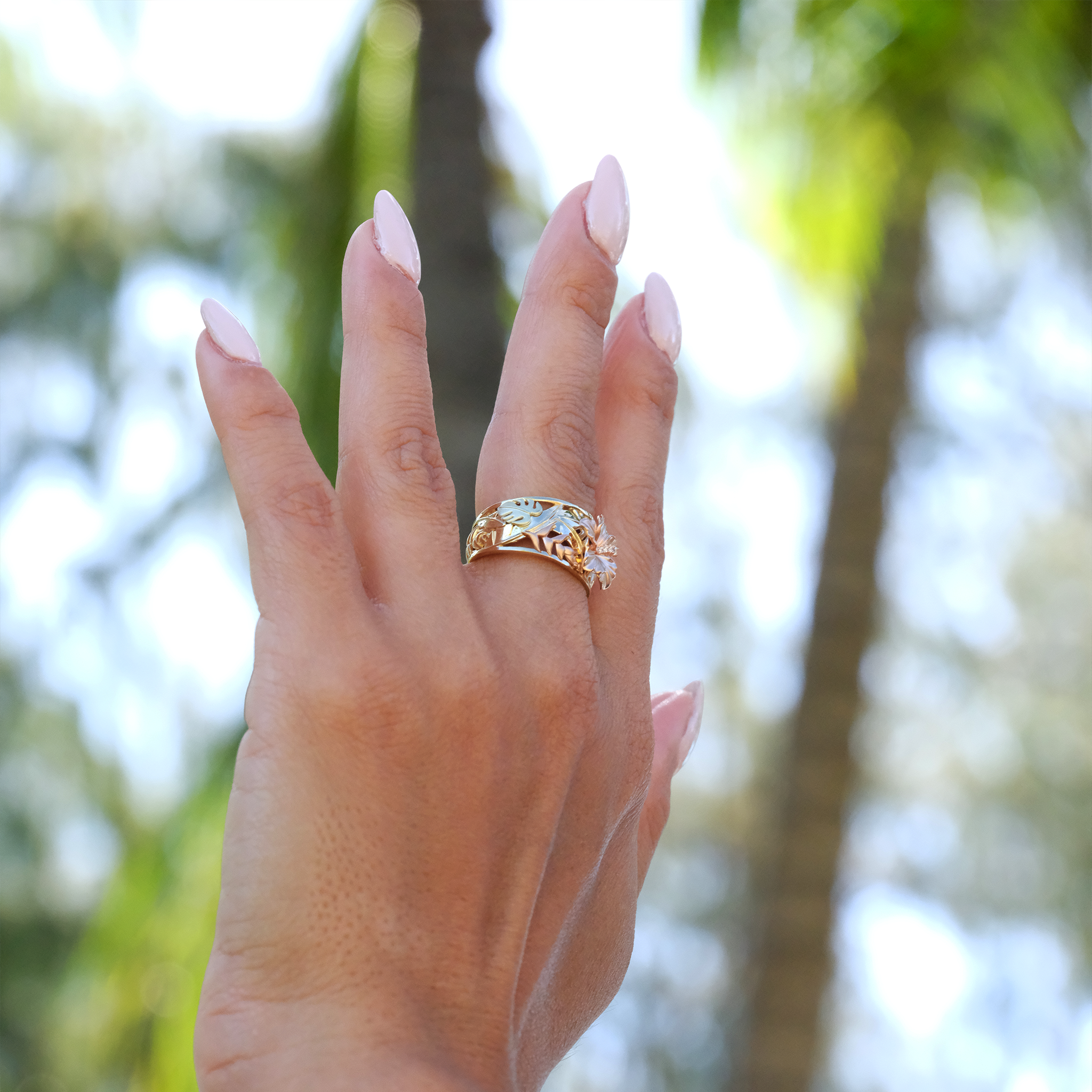 A woman's hand with a Hawaiian Gardens Hibiscus Ring in Tri Color Gold with Diamonds against blurred background