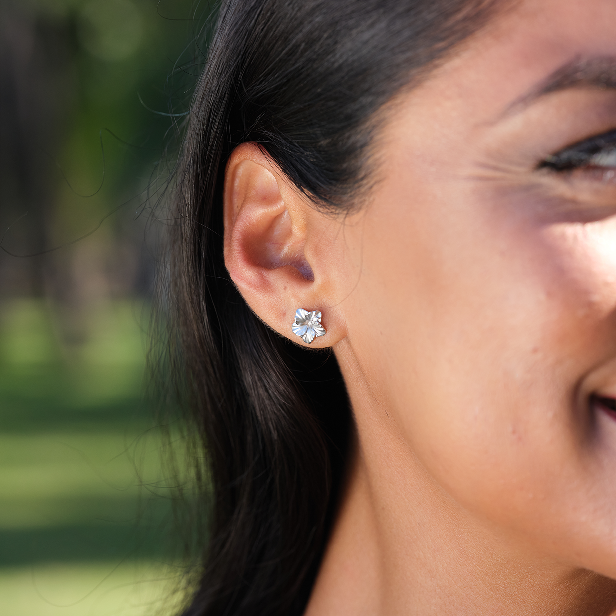 Close up of 9.5mm Diamond Hibiscus Earrings in 14k White Gold on ear with blurred green background
