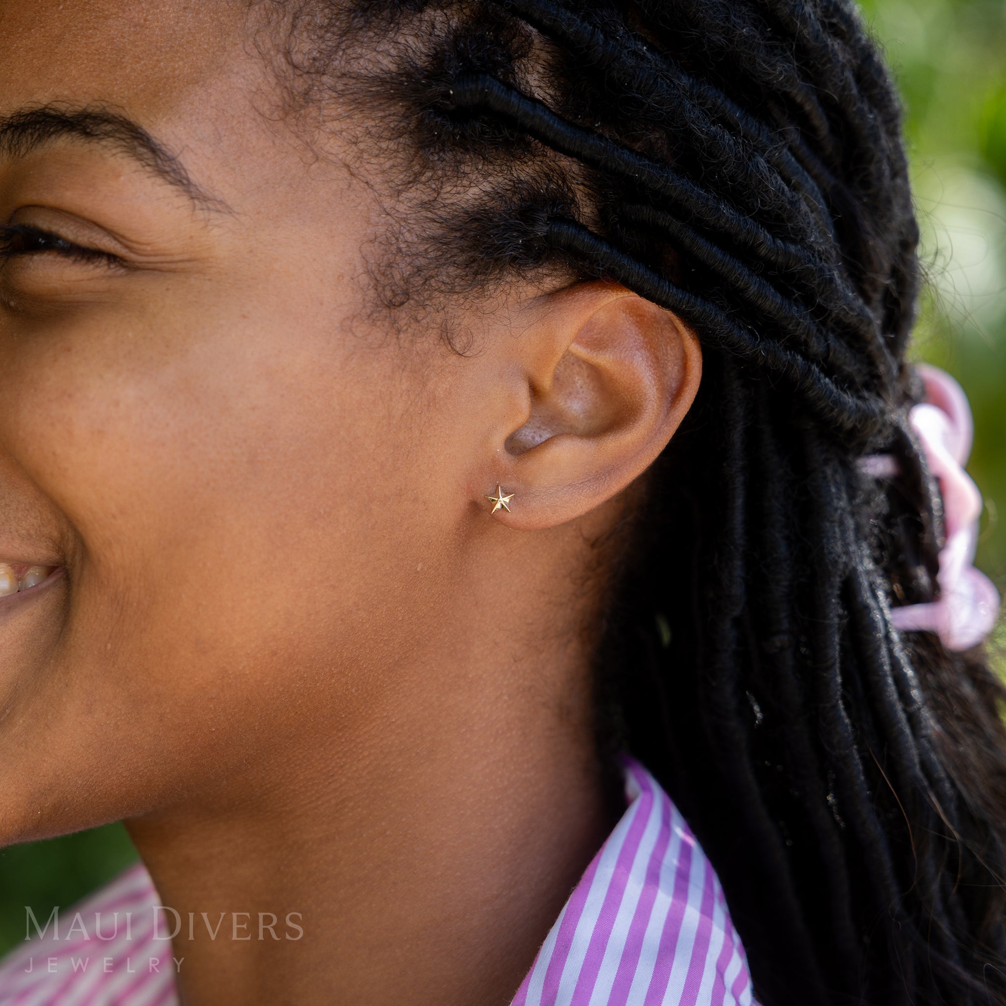 A side view of a smiling woman in a pink and white striped top wearing a Starfish Earring in 14k yellow gold, against a blurred leafy green background