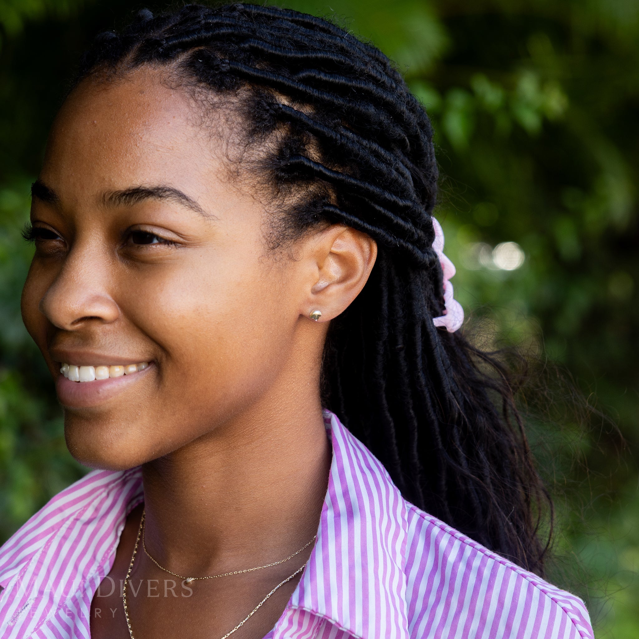 A side view of a smiling woman in a pink and white striped top wearing a Scallop Shell Earring in 14k yellow gold, paired with a tiny diamond heart necklace, against a blurred leafy green background