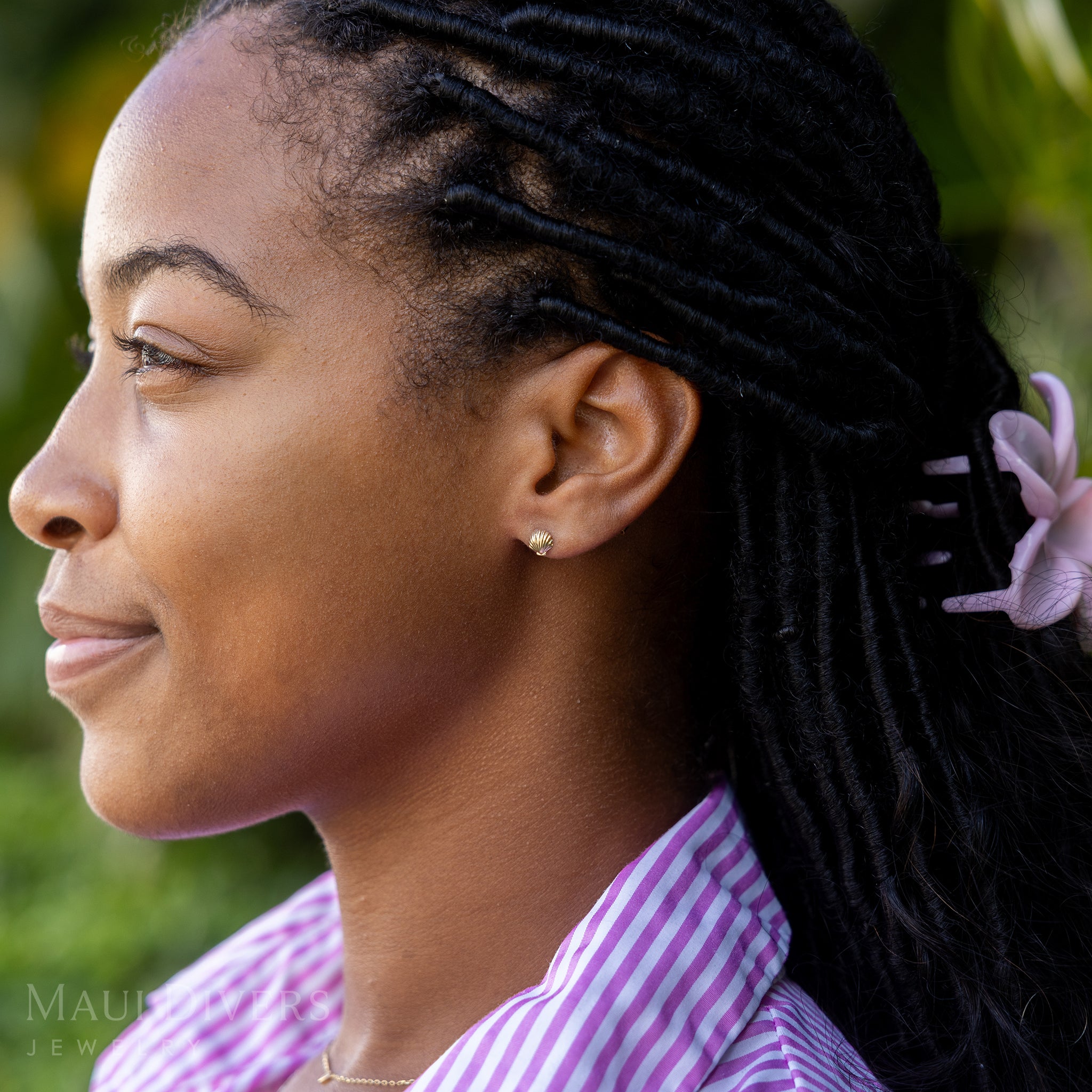 A side view of a smiling woman in a pink and white stripe top wearing a Hawaiian Sunrise Shell Earring in 14k yellow gold, against a blurred leafy green background