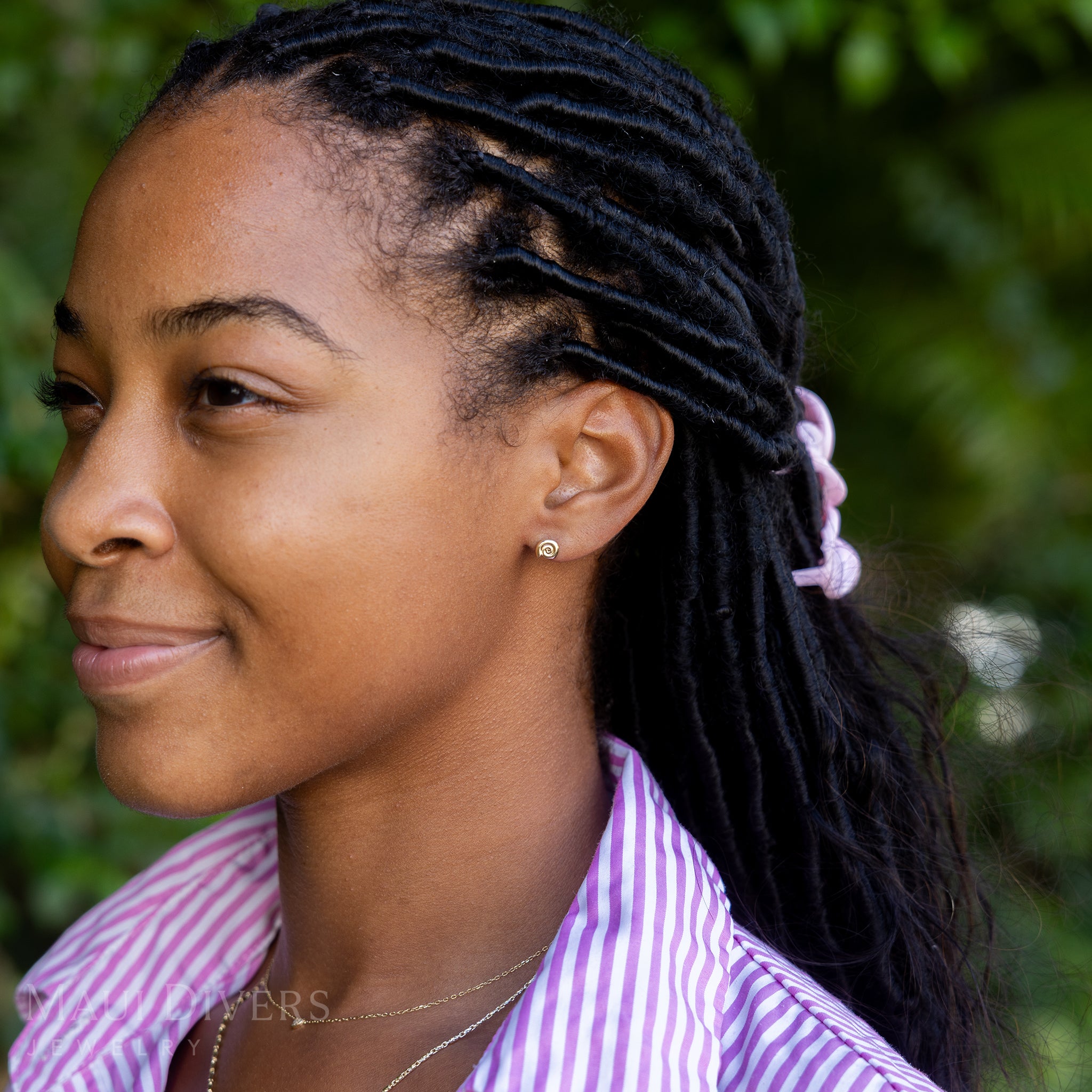 A side view of a smiling woman in a pink and white stripe top wearing a Conch Shell Earring in 14k yellow gold, paired with a tiny diamond necklace, against a blurred leafy green background