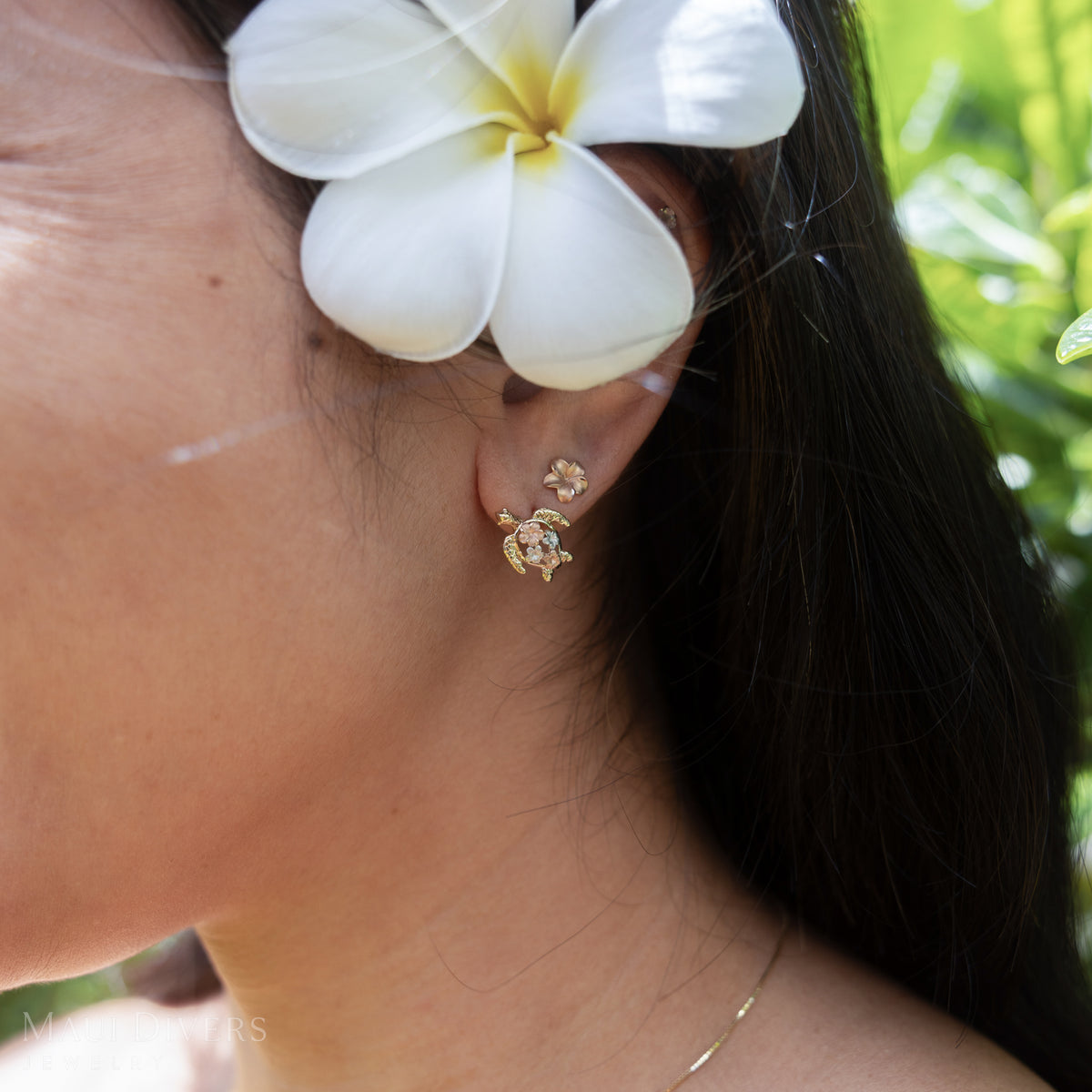 Close-up of a womanʻs ear with a plumeria behind it and wearing a sea turtle earring with white and rose gold plumeria and a small rose gold plumeria earring against a blurry nature background