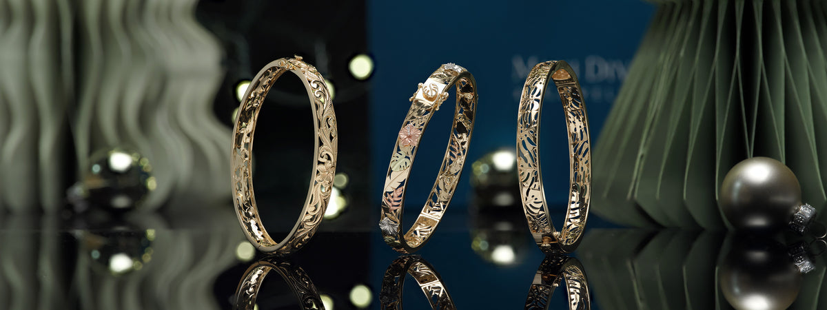 Three ornate gold bangles with intricate Hawaiian designs are displayed on a reflective black surface against a dark blue and green background with decorative elements.