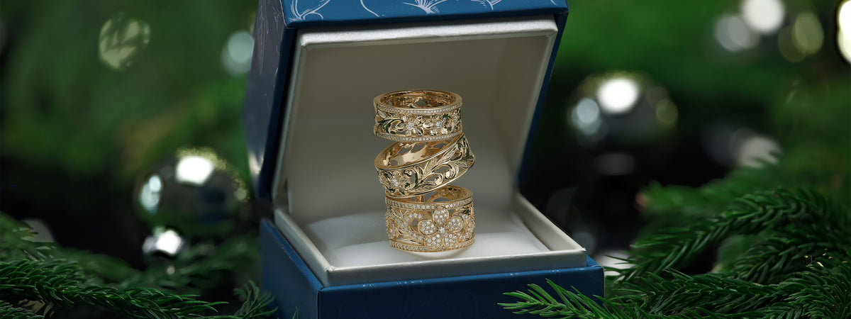 Three gold Hawaiian Heirloom rings with intricate diamond-set patterns displayed inside an open blue jewelry box, surrounded by green pine branches and festive bokeh lights.