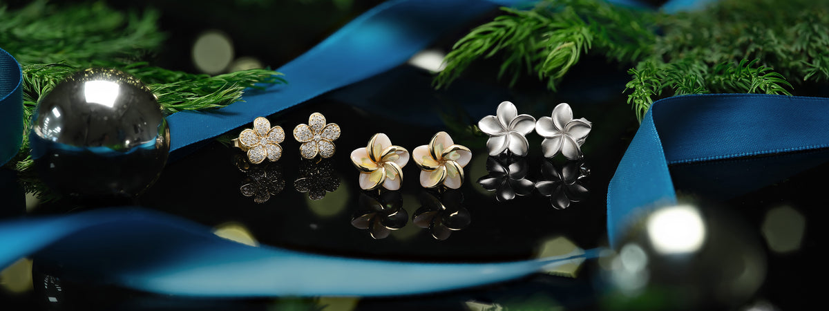 Three plumeria flower-shaped stud earrings arranged on a glossy black surface with blue ribbon, green foliage, and reflective holiday ornaments in the background.