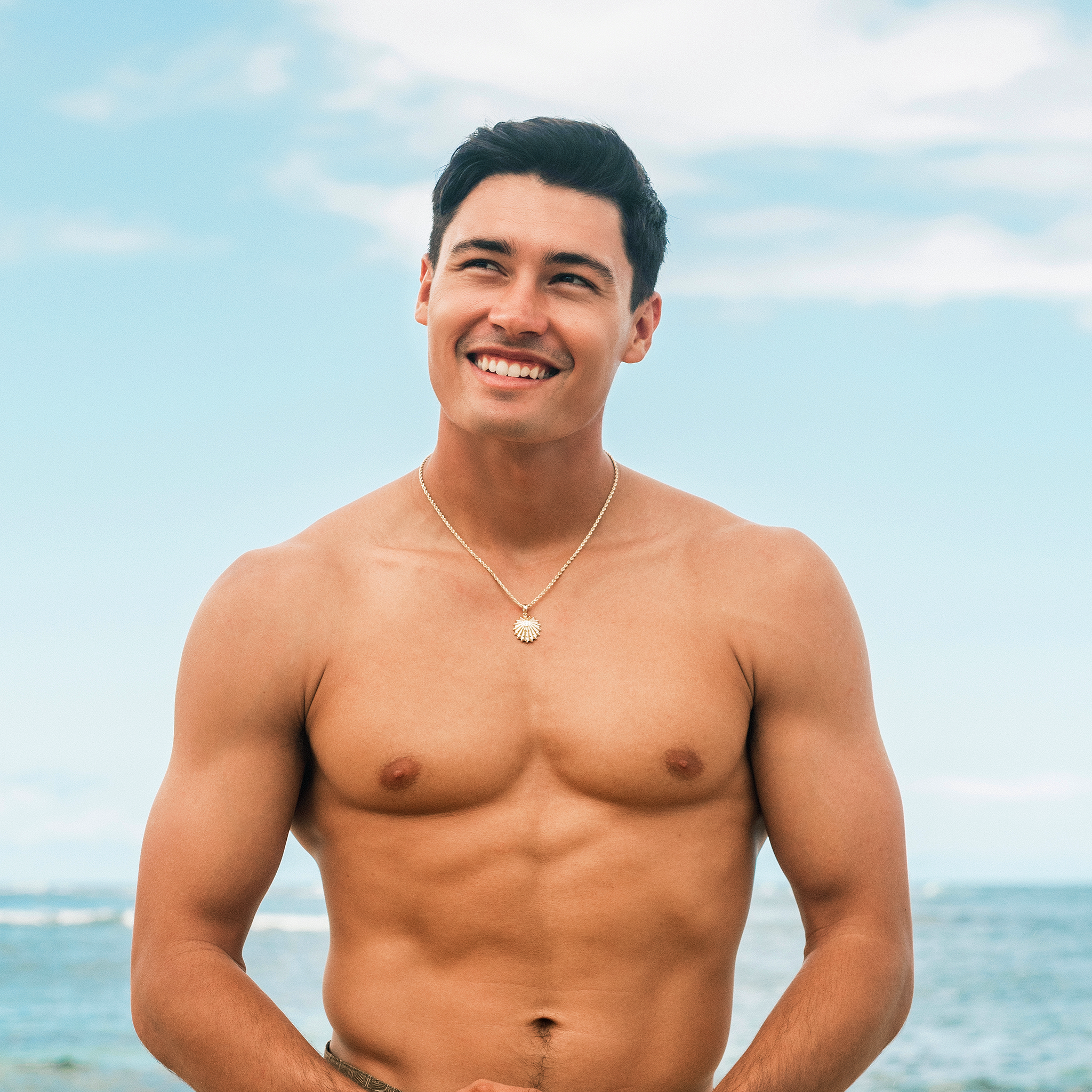 Man with an ʻOpihi Pendant in Gold with Lab Grown Diamond standing on a beach with clear blue sky and ocean.
