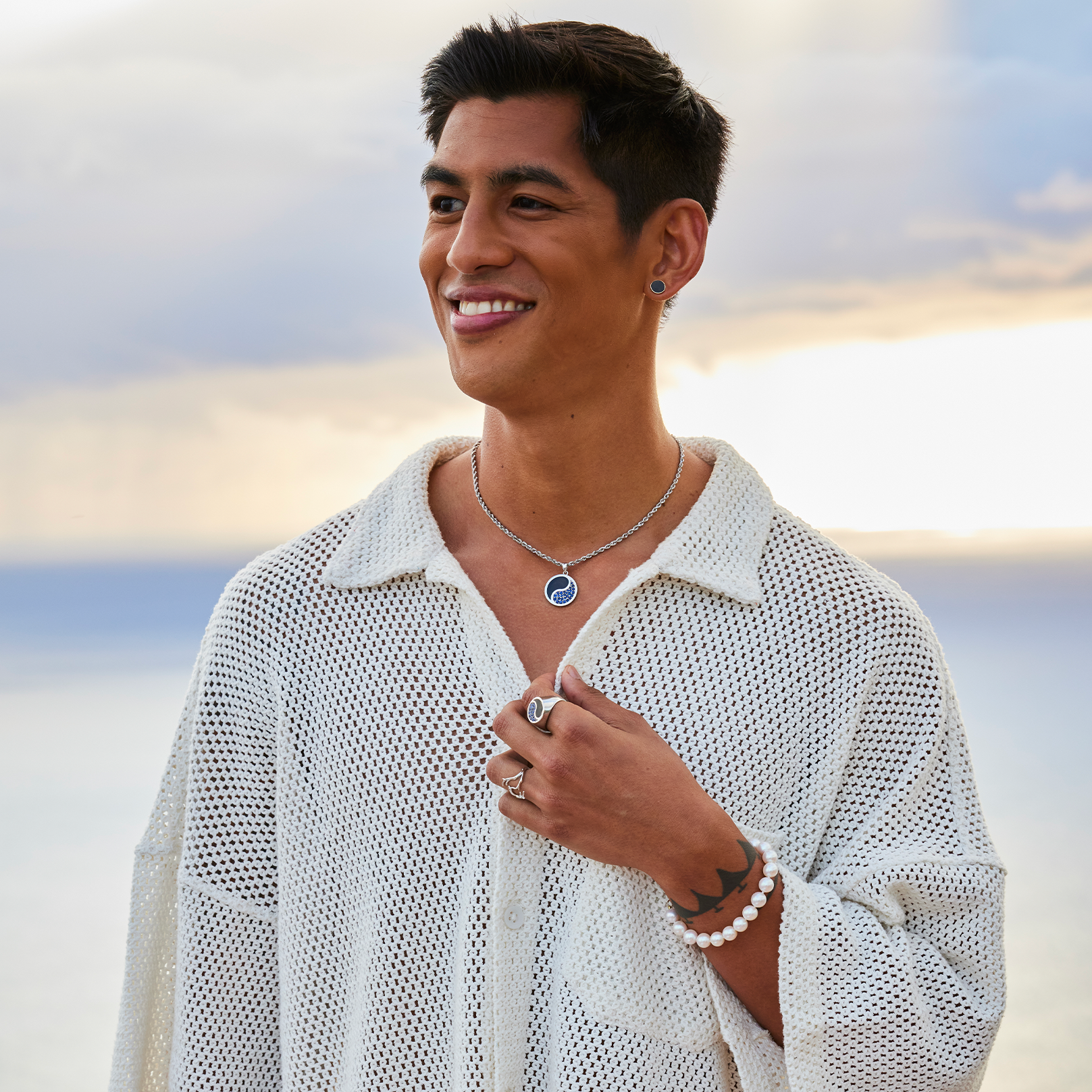 Smiling man wearing a freshwater white pearl bracelet, with 14k white gold rings featuring coral, blue sapphires, and Hawaiian black coral, plus a coordinating sapphire and black coral necklace and black coral earrings