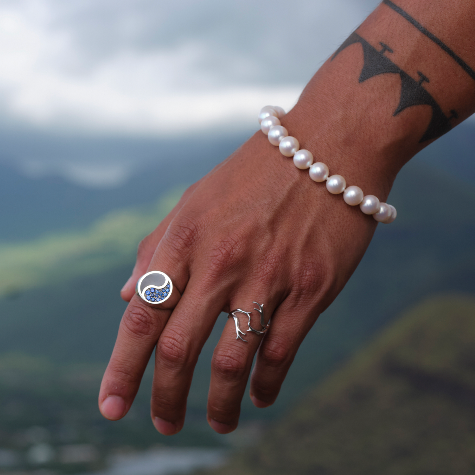 Close-up of a man wearing a freshwater white pearl bracelet and two 14k white gold rings, one featuring a coral design and the other set with blue sapphires and Hawaiian black coral