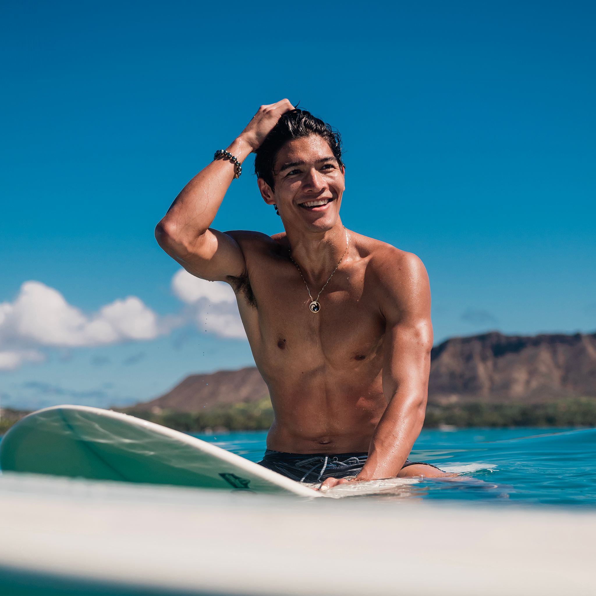 Man on a surfboard in the ocean wearing a 2.7mm Paperclip Chain in 14k Yellow Gold and wave pendant