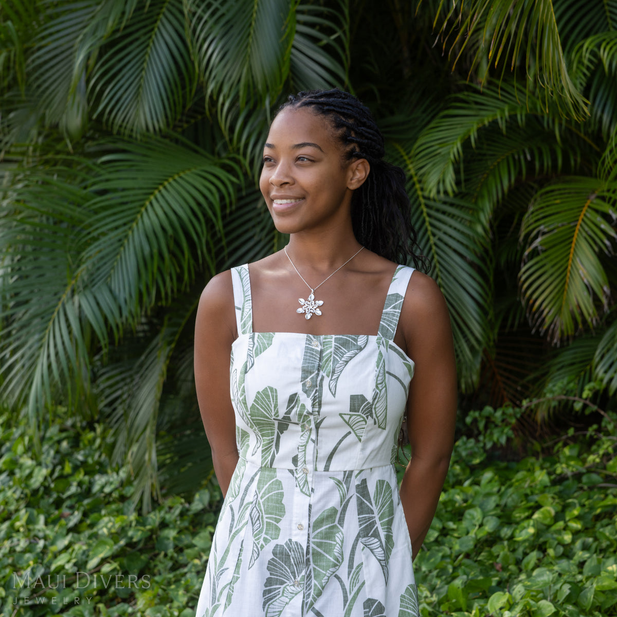A woman wearing a white sleeveless dress with a green tropical leaf print stands outdoors in front of dense green palm foliage. She is wearing a silver kalo snowflake pendant necklace.