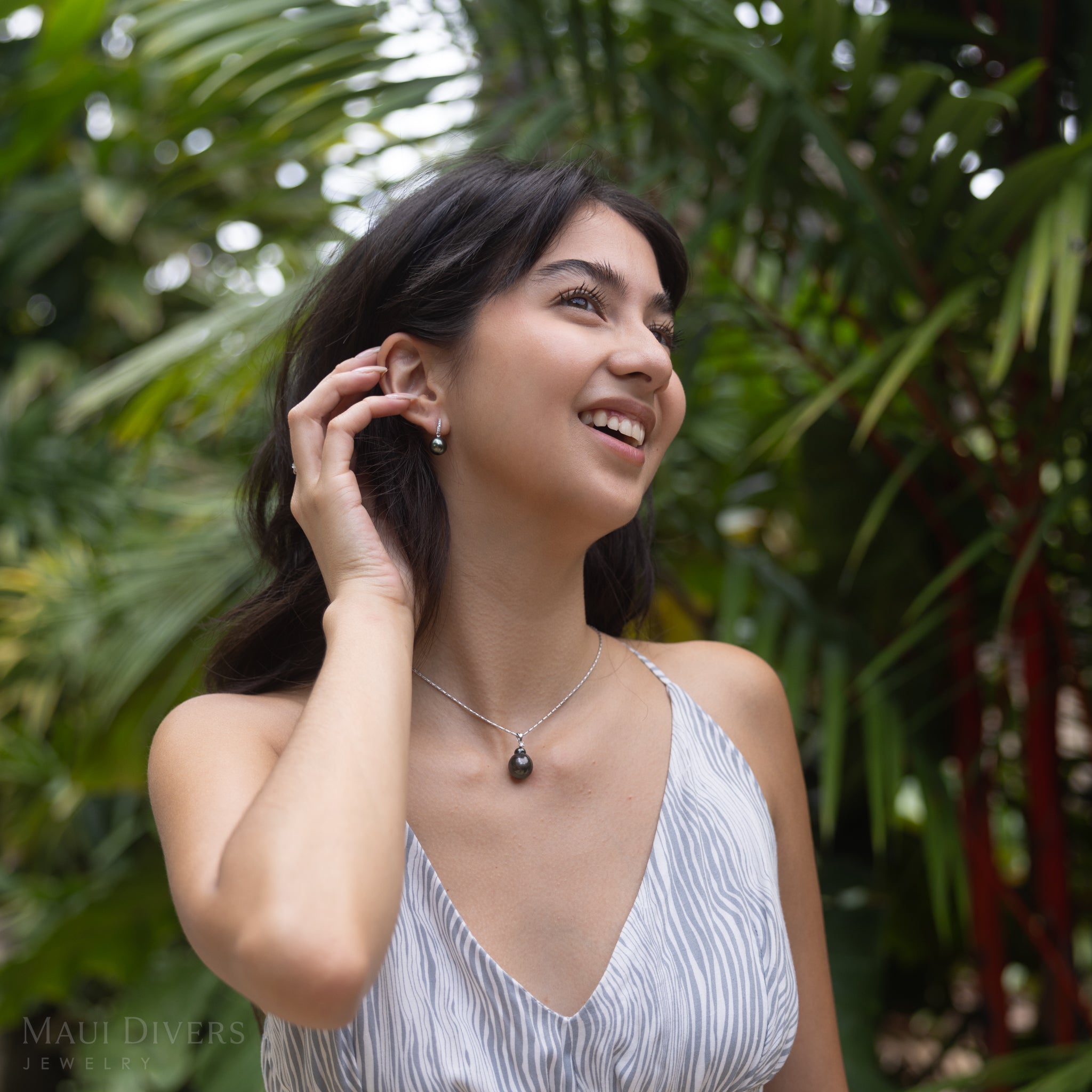 Tahitian black pearl earrings and pendant in white gold worn by a smiling woman in a tropical forest.