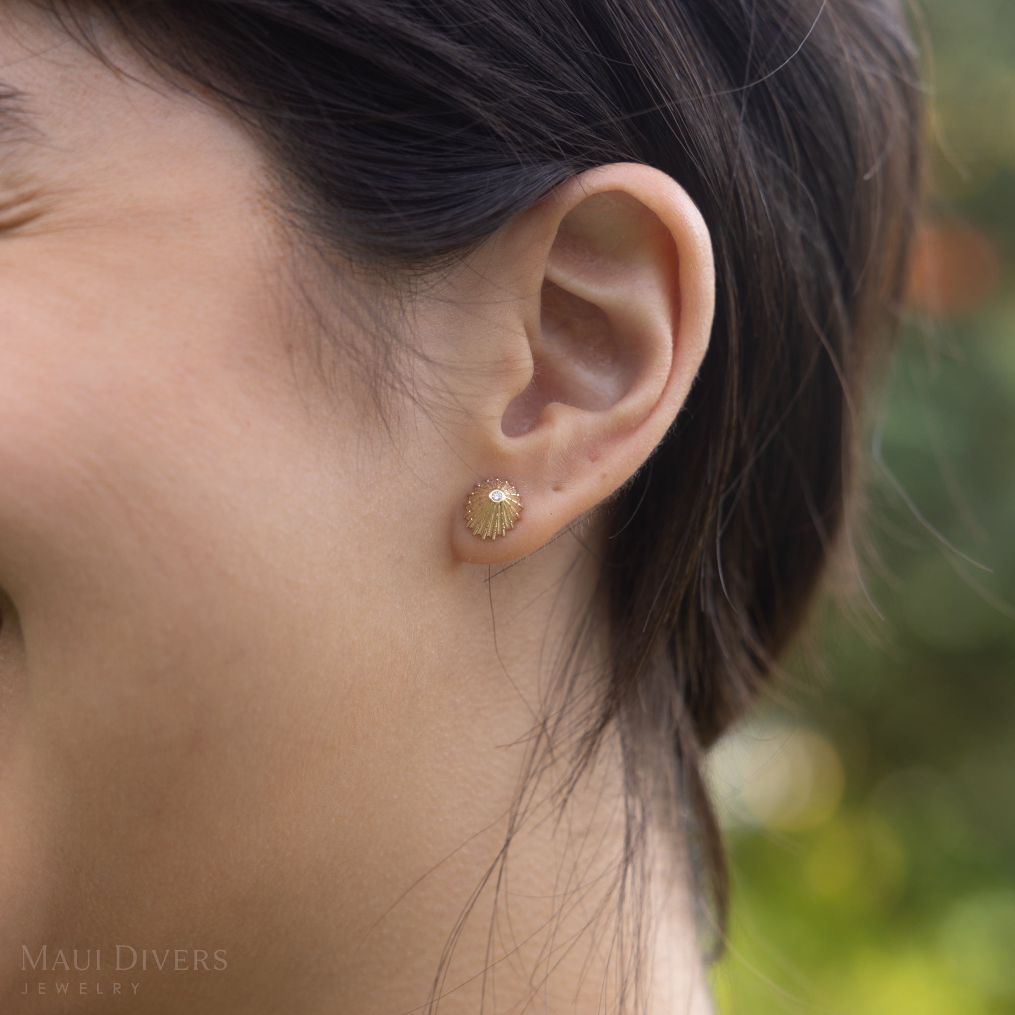 Close up of 10mm ʻOpihi Earrings in Gold with Diamonds on ear with blurred green background
