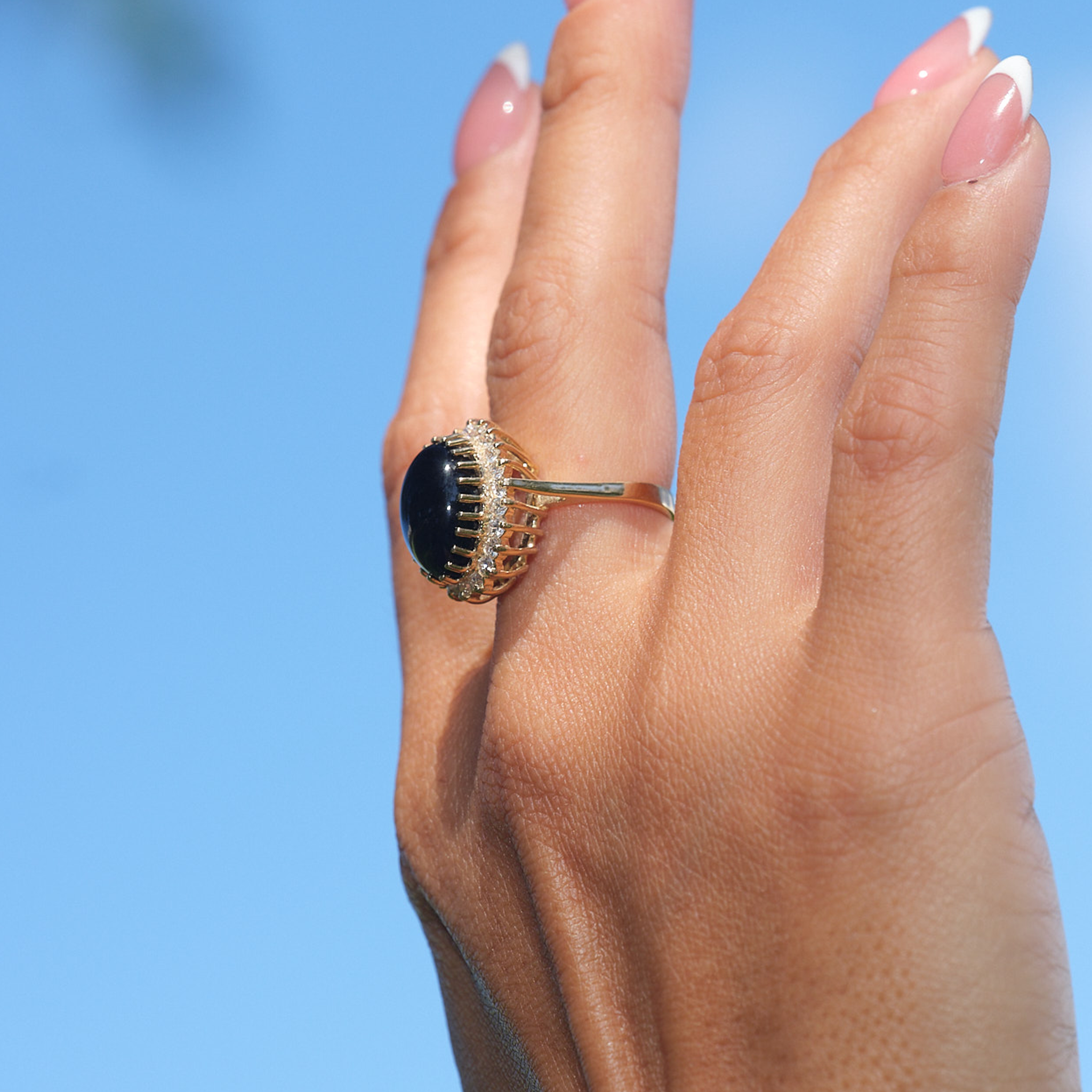 A woman's hand wearing a Princess Ka‘iulani Black Coral Ring in Gold with Diamonds against blue sky