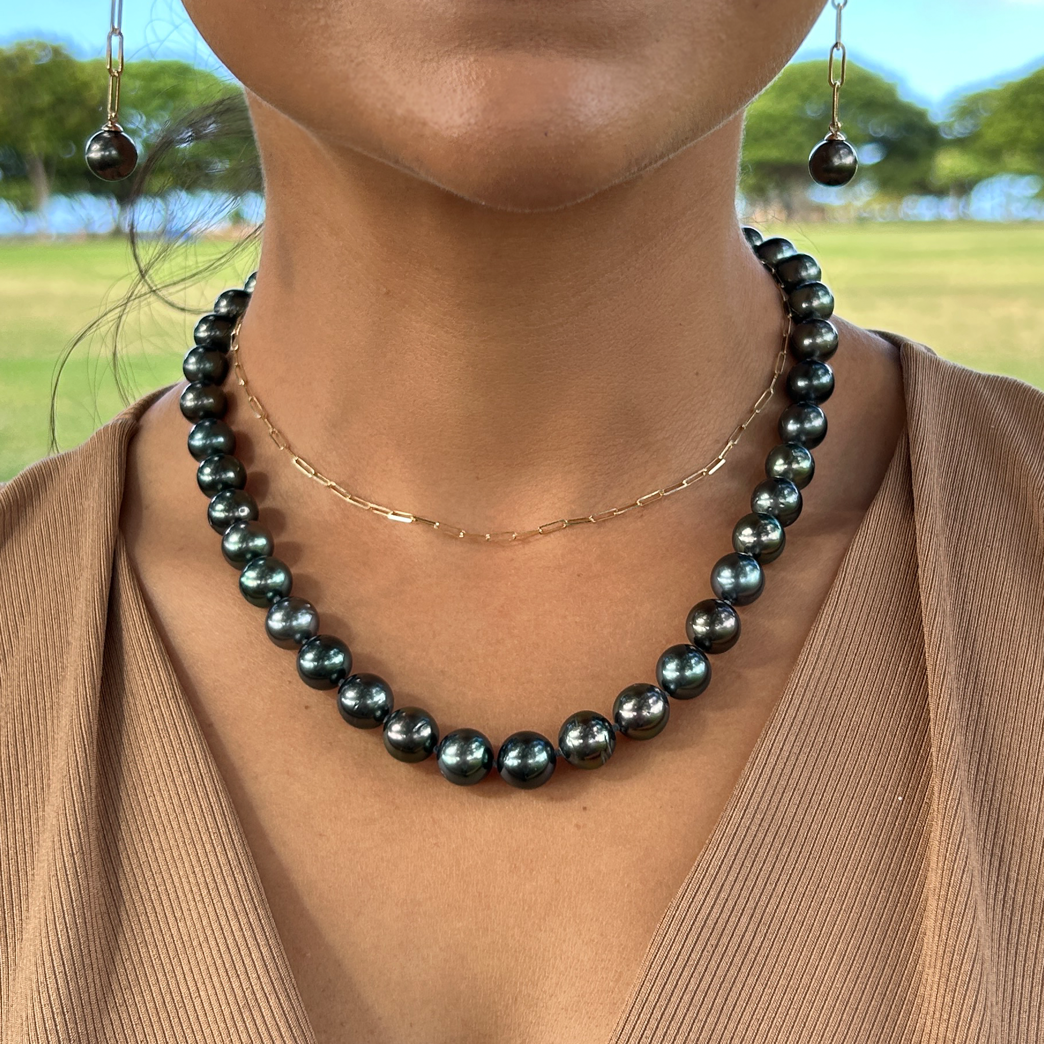 Close up of a woman wearing a Tahitian black pearl strand and paperclip chain necklace with a blurry green field background
