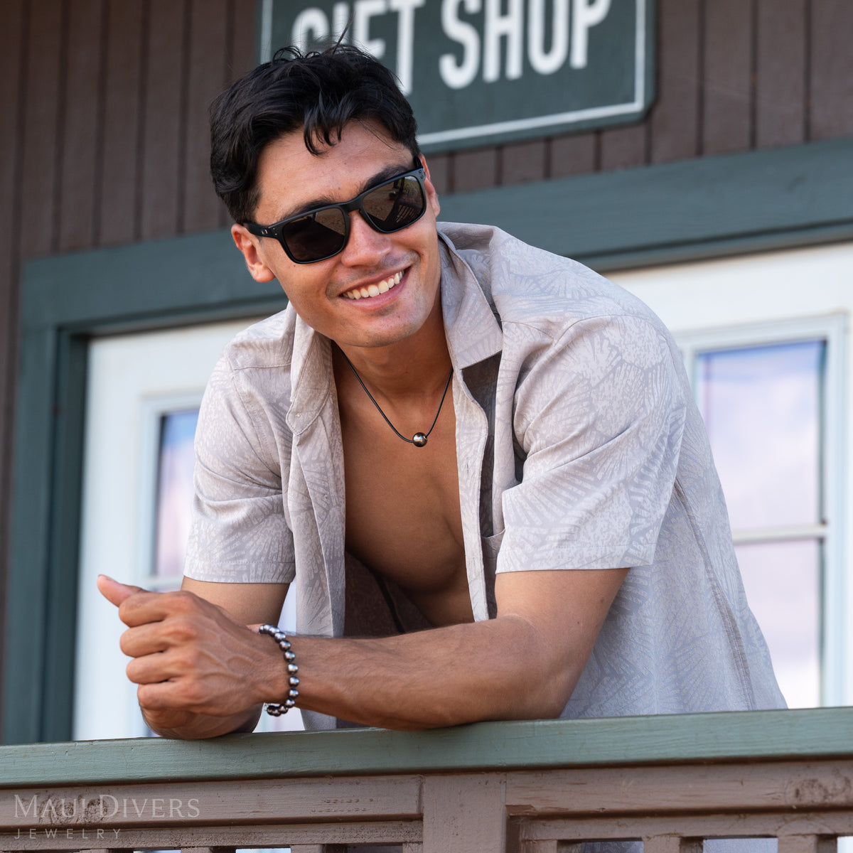  A smiling man in glasses and a patterned grey shirt wearing a black coral Tahitian black pearl necklace and matching pearl bracelet, leaning on a wooden rail