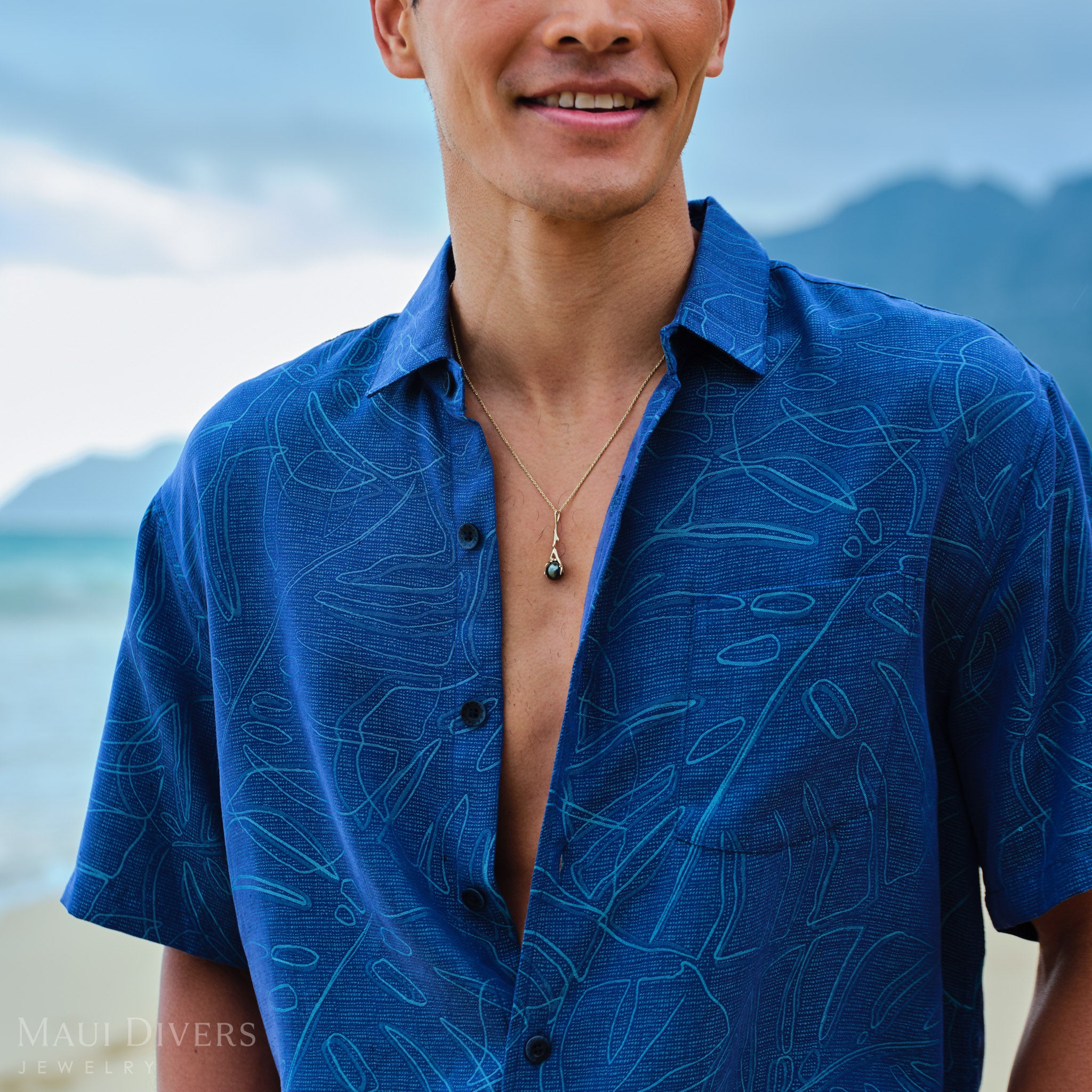 Man wearing gold Tahitian black pearl pendant a blue patterned shirt with a beach and mountains in the background