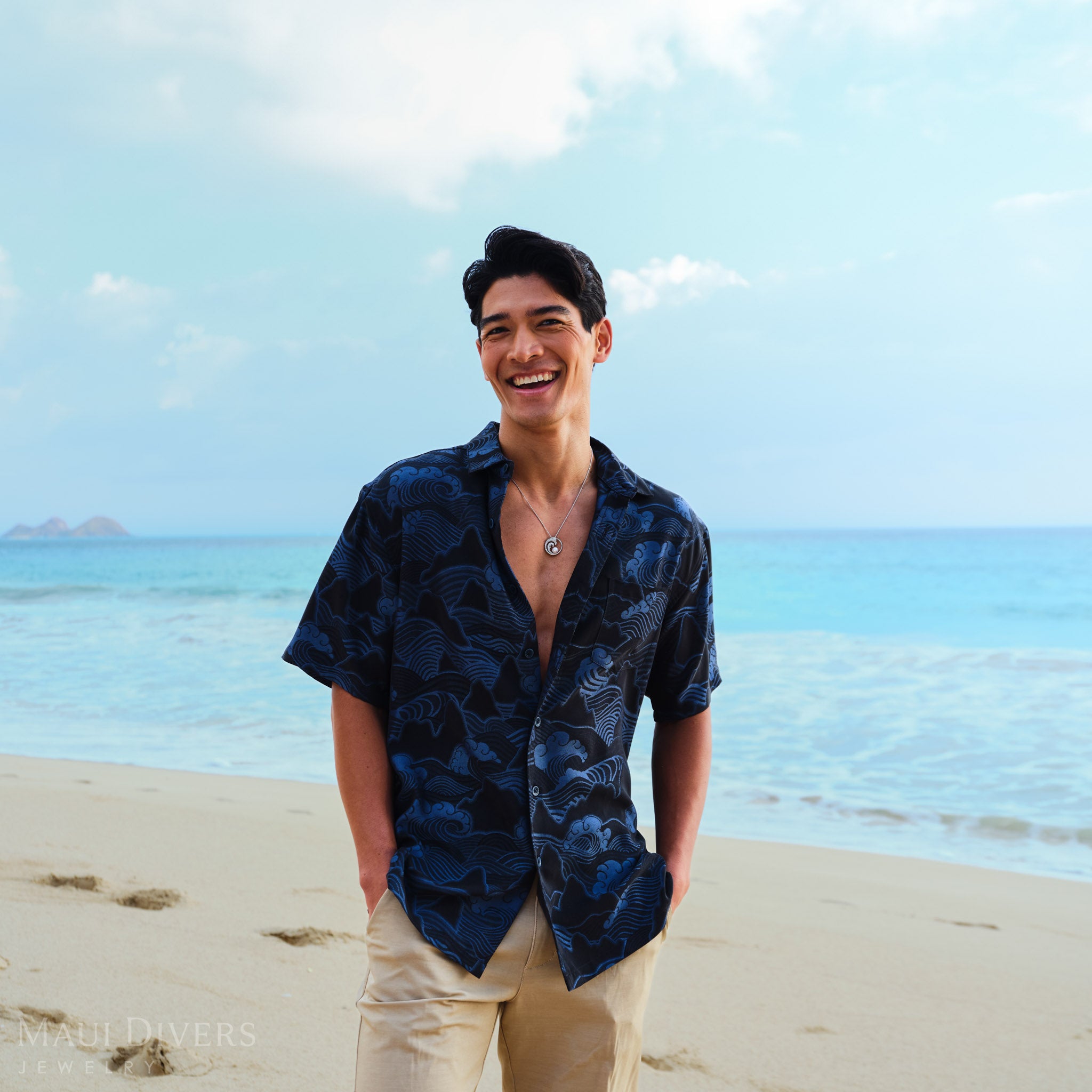 Man in a blue patterned shirt wearing a white gold wave pendant with an Akoya pearl standing on a beach with clear blue water and sky