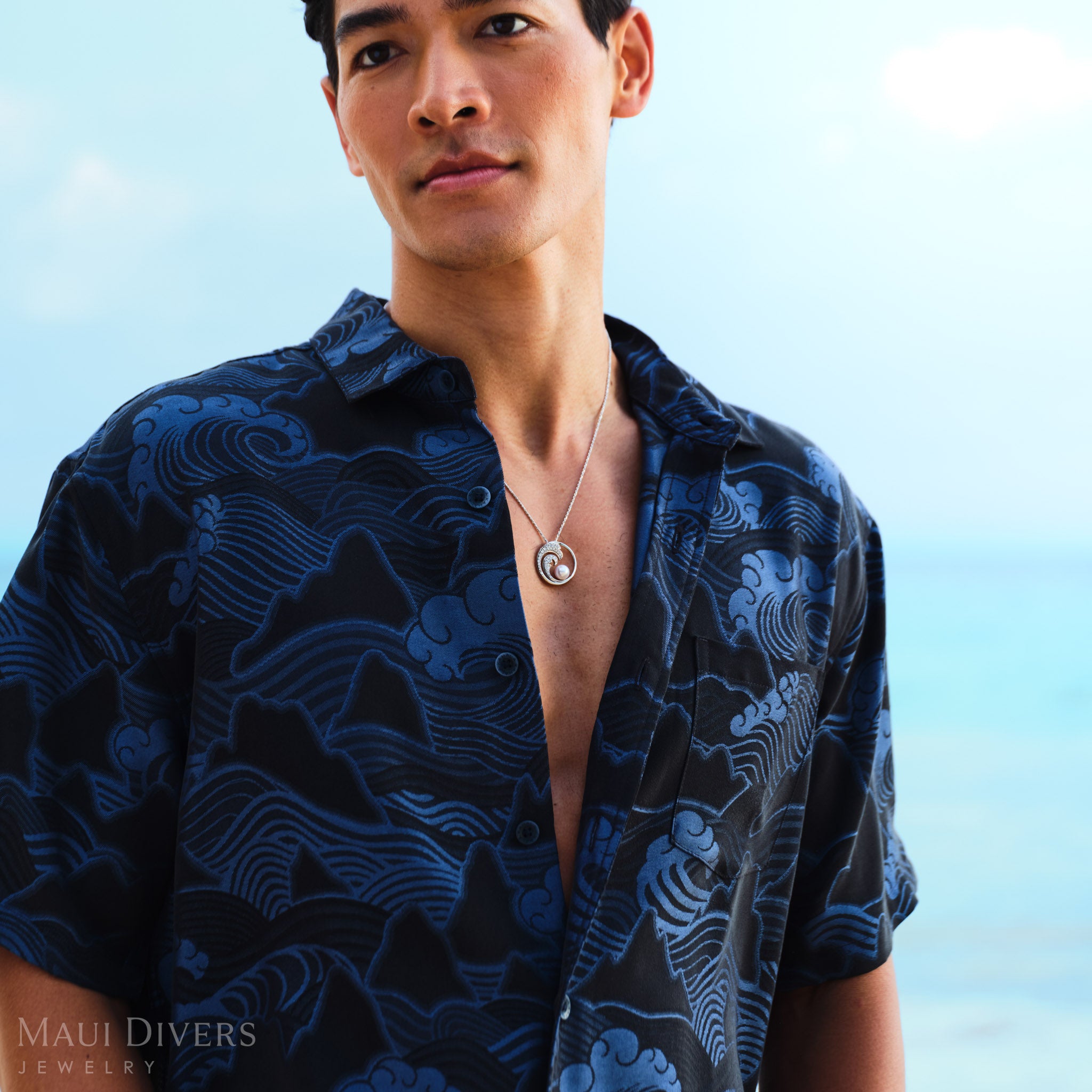 Man wearing a blue patterned shirt with a Nalu Akoya white pearl pendant in white gold, standing against a clear sky.