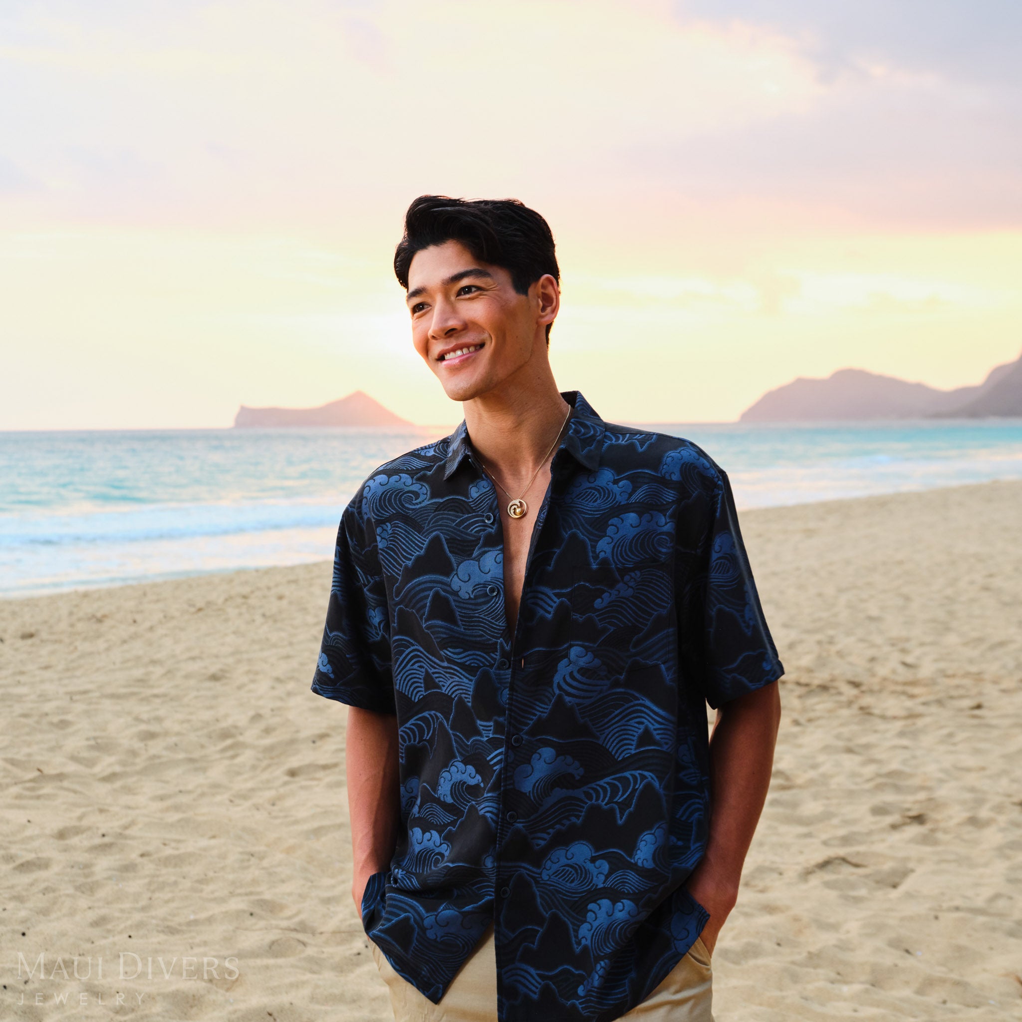 Man wearing a blue patterned shirt wearing a gold diamond wave South Sea gold pearl pendant on a beach with a sunset in the background
