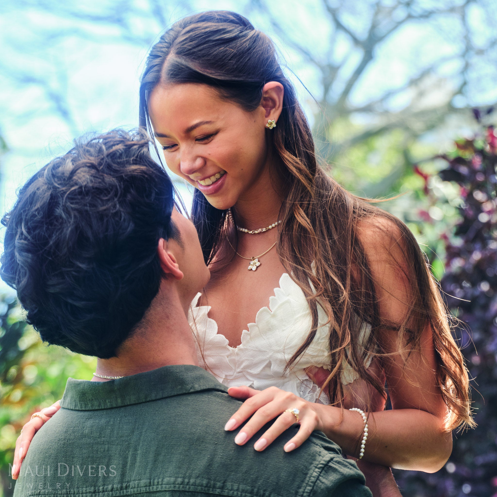 Smiling woman in a white top wearing a Plumeria Mother of Pearl Pendant and Earring in 14k yellow gold with a Hawaiian Heirloom freshwater pearl strand necklace, being lifted by a smiling man against a blurred leafy green background