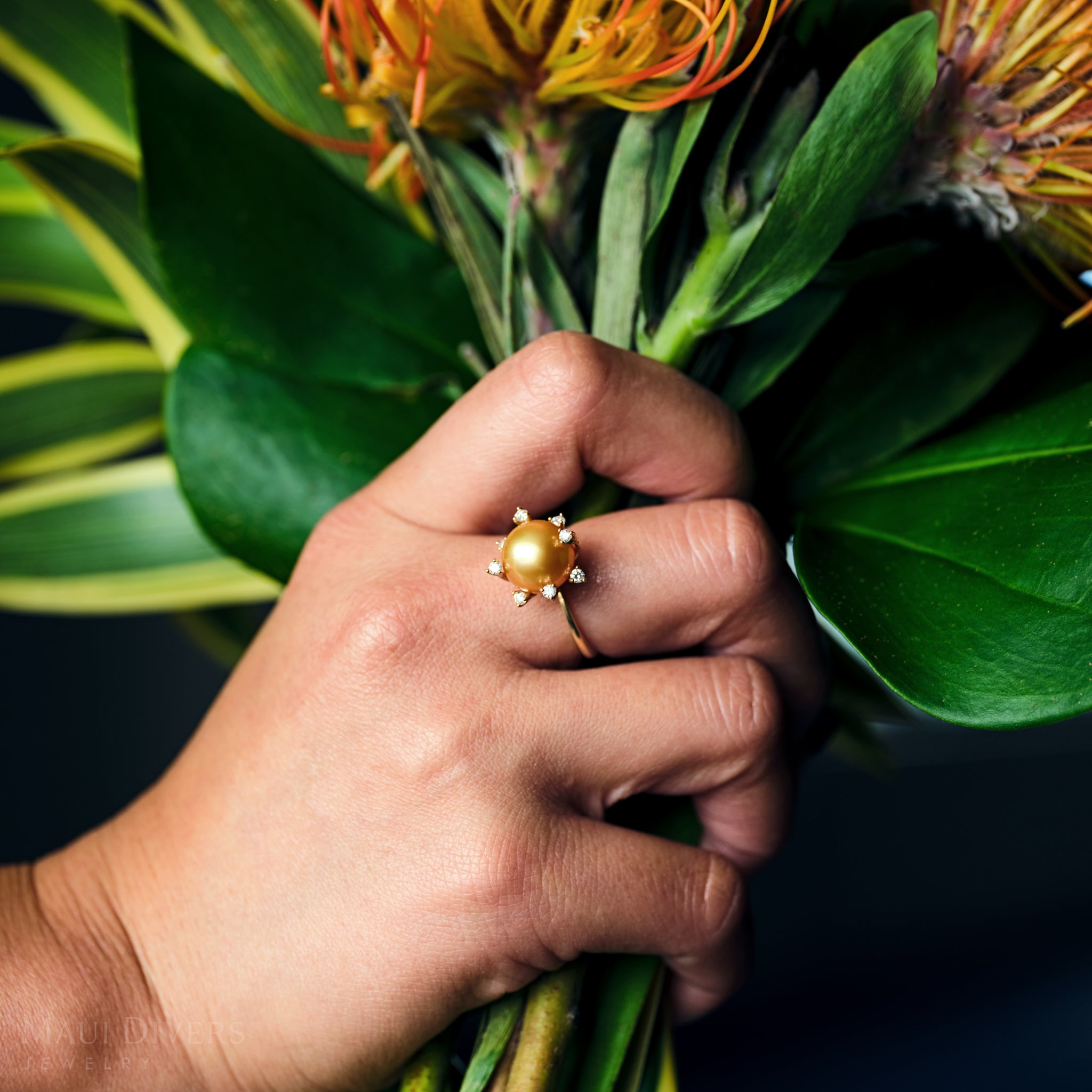 Close-up of a South Sea gold pearl ring with diamonds in 14k yellow gold worn on a hand holding a bouquet of flowers.