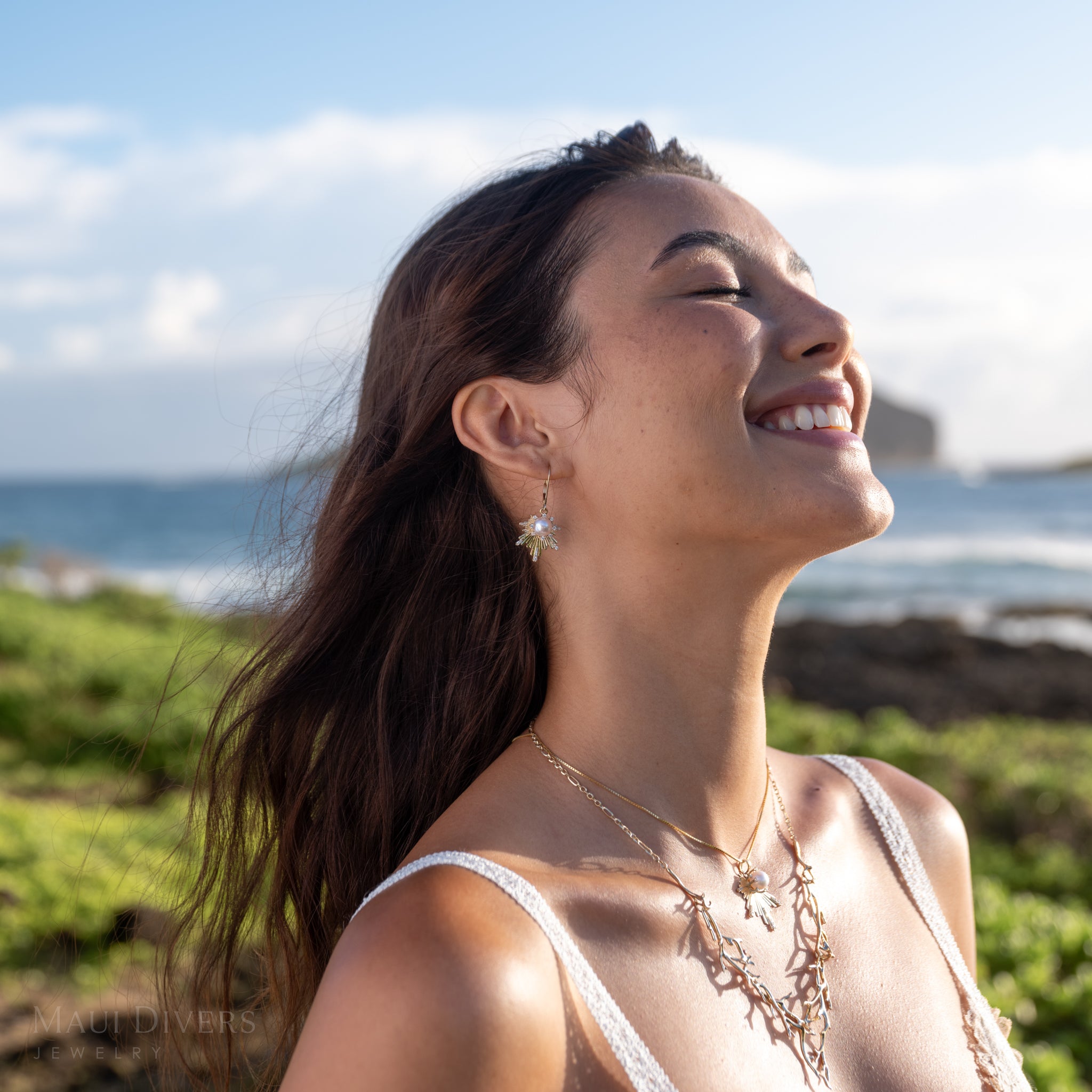 Woman wearing the E Hoʻāla Akoya White Pearl and diamond earrings in yellow gold and a matching pendant, smiling outdoors with the ocean in the background