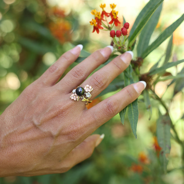 Womanʻs hand wearing Pearls in Bloom Plumeria Tahitian Black Pearl Ring in Tri Color Gold with Diamonds - 22mm as she touches a red flower