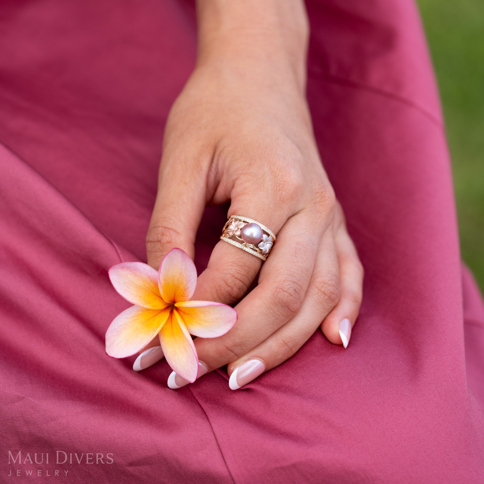 Close up of a hand wearing a Natural Lavender Freshwater Pearl Ring with Tri Color Plumerias in 14k Gold accented with  Diamonds holding a light pink and yellow plumeria flower.