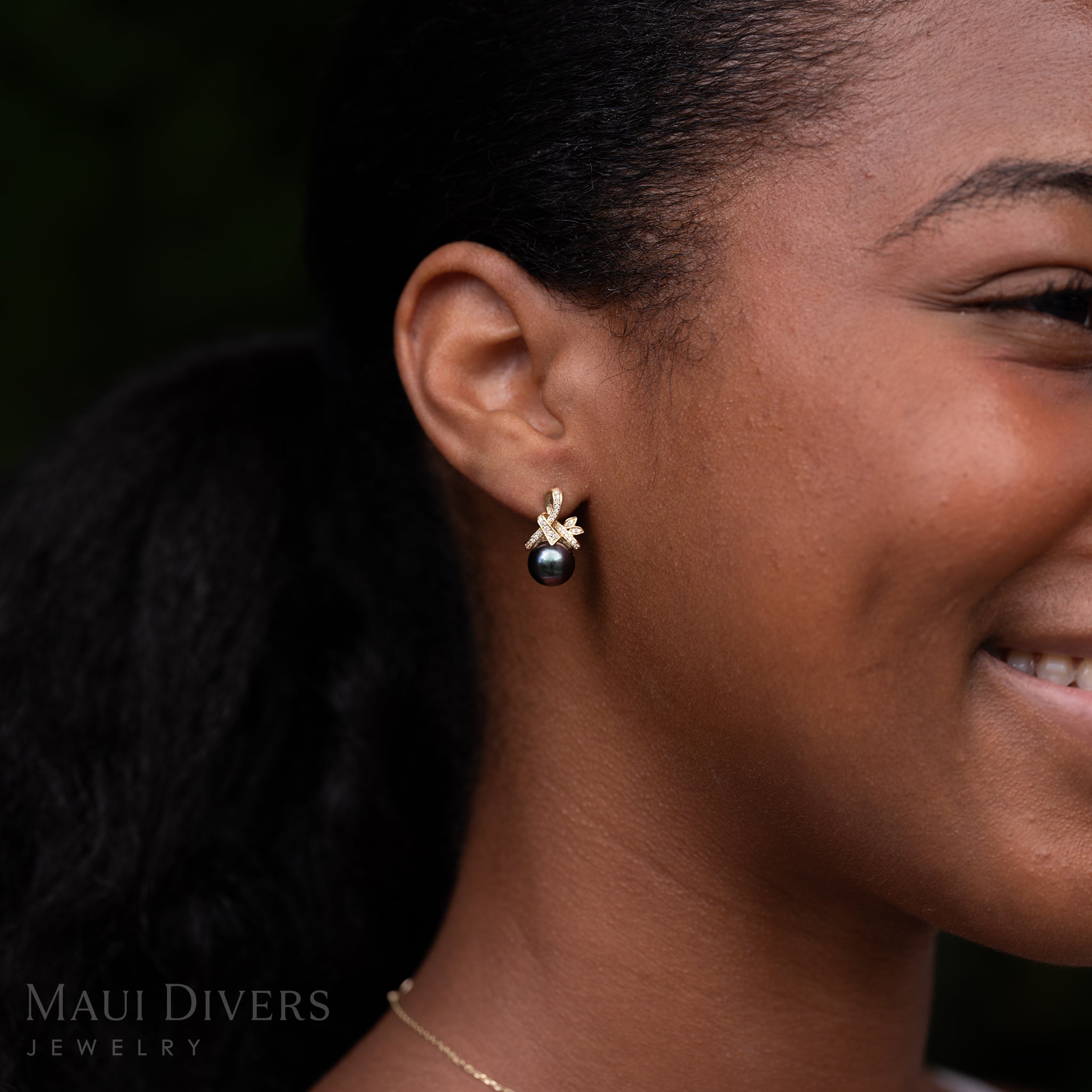 Close-up of a smiling woman wearing a Tahitian Black Pearl Earring in 14k Yellow Gold with Diamonds on her ear against a blurred dark background