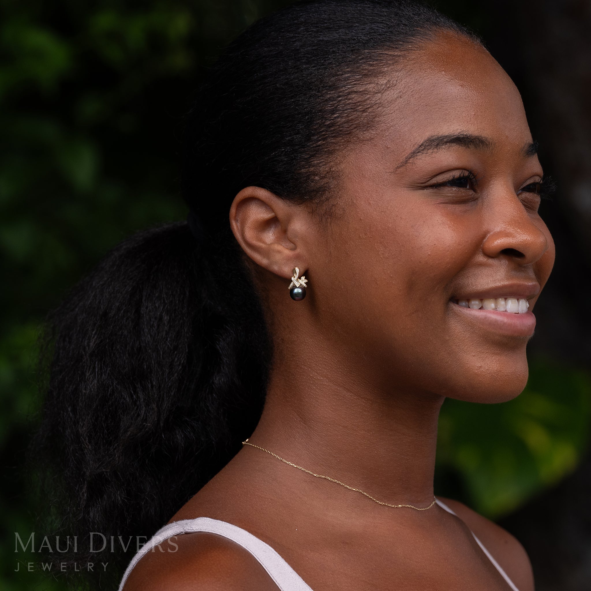 A smiling woman wearing a Tahitian Black Pearl Earring in 14k Yellow Gold with Diamonds on her ear against a blurred leafy background