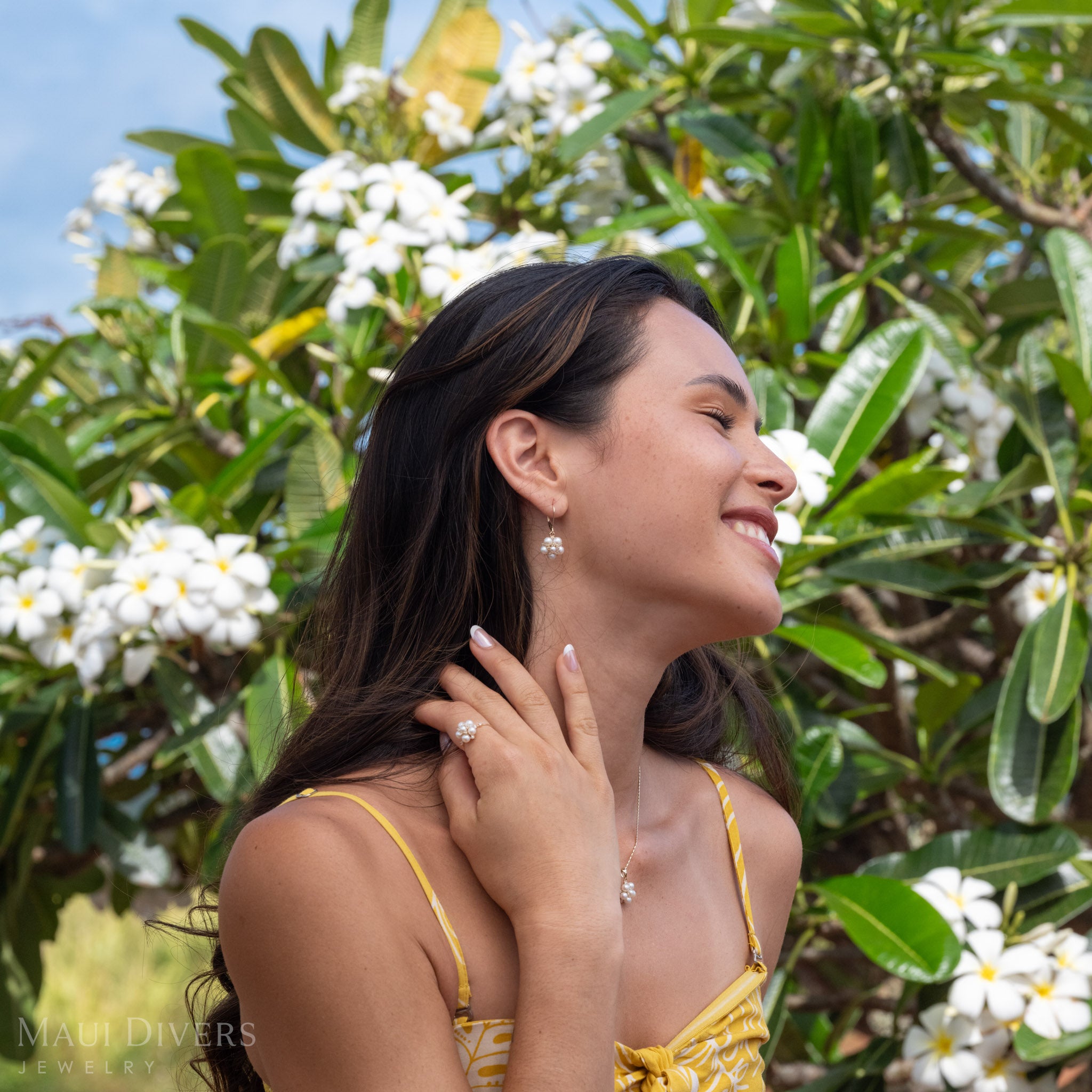 Woman wearing Tiny Natural White Freshwater Pearls on a 14k Gold Ring with Diamonds and the matching earring and necklace set in front of a blurred plumeria tree background