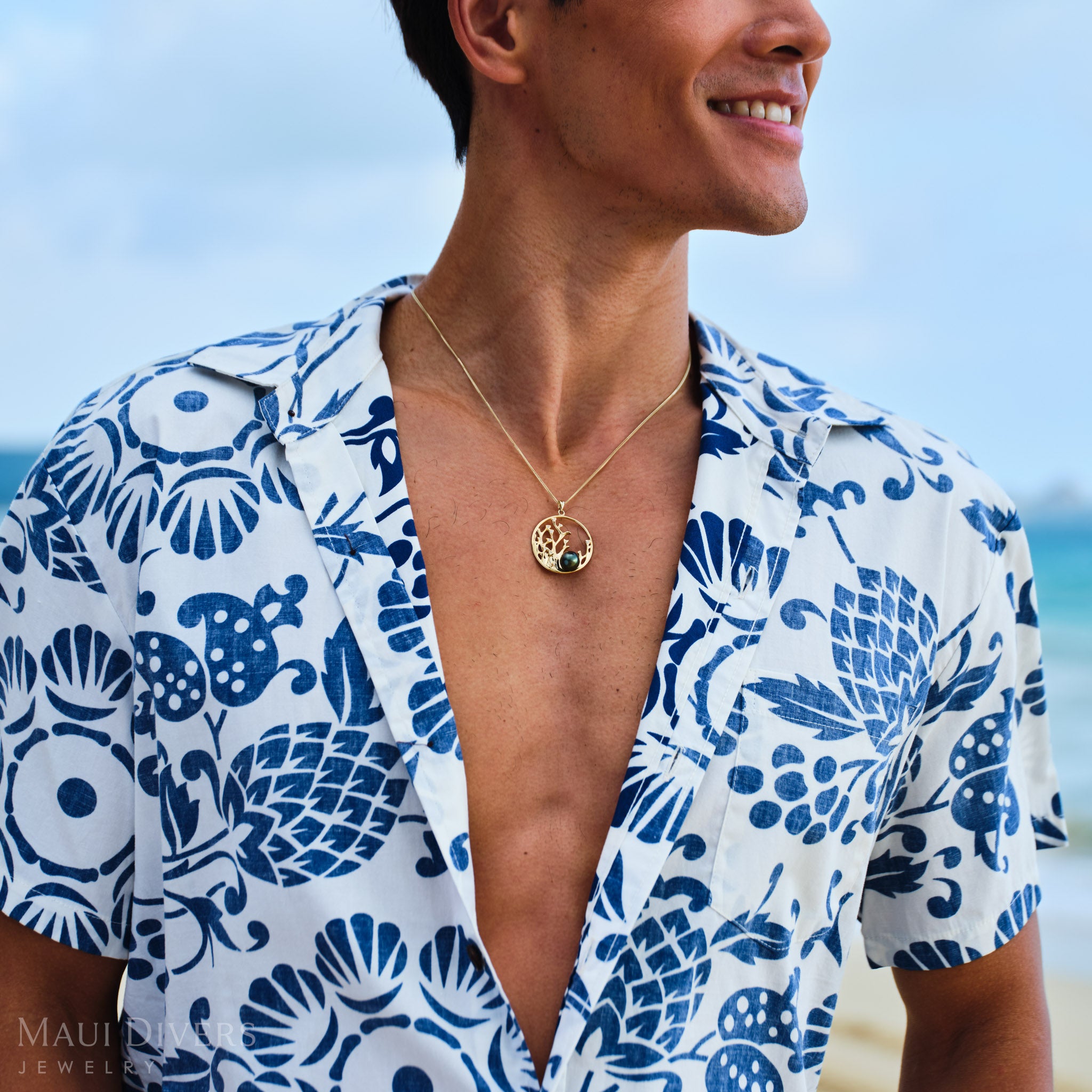 Man wearing a blue and white patterned shirt with a Reefs Tahitian Black Pearl Pendant in Gold on a beach.