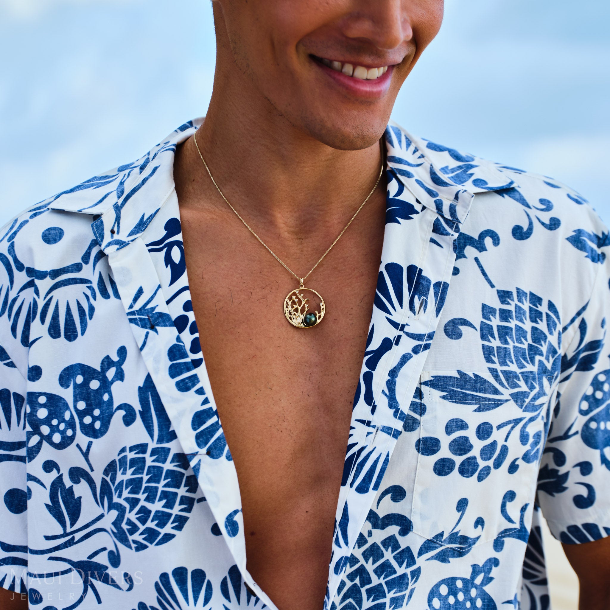 Person wearing a blue and white patterned shirt with a Reefs Tahitian Black Pearl Pendant in Gold against a clear sky.