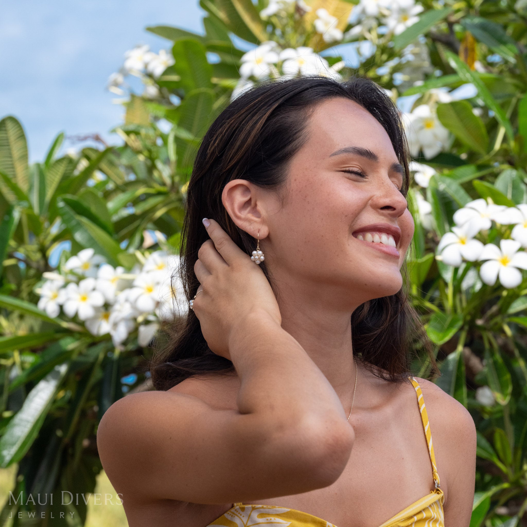 Woman smiling wearing Tiny Natural Freshwater White Pearls on a 14k Gold earring accented with Diamonds, her hand in her hair in front of a blurred green and white plumeria background. 
