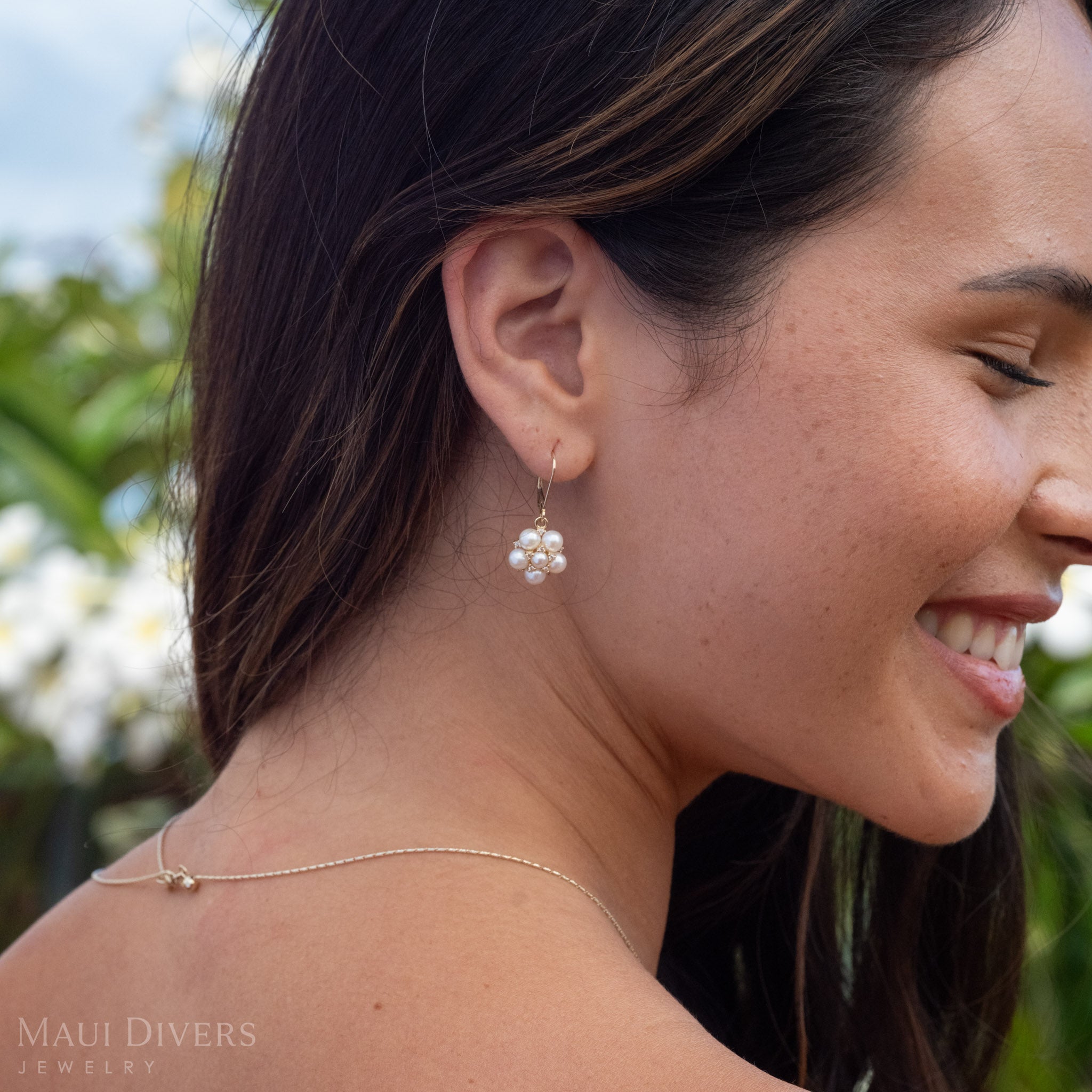 Close up side profile of a woman wearing Tiny Natural Freshwater White Pearls on a 14k Gold earring accented with Diamonds with a blurred green and white plumeria background 