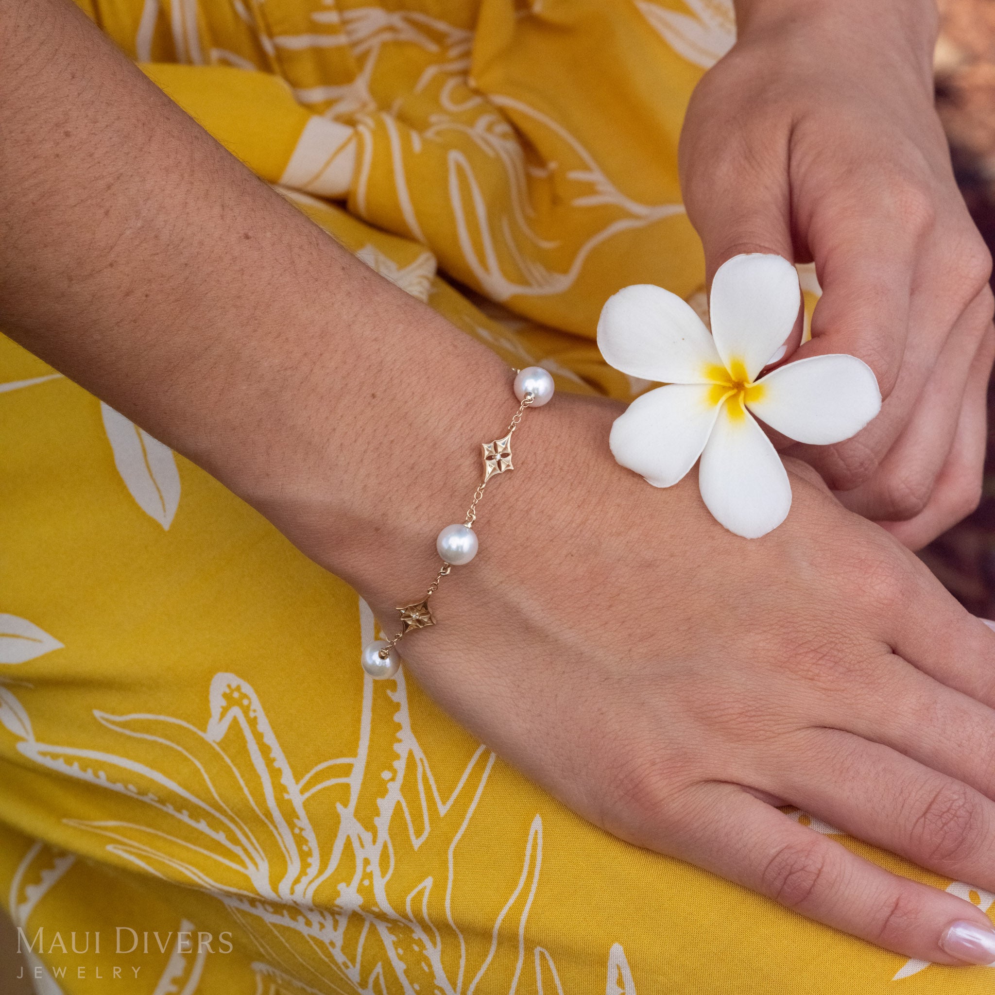 Close up of a 14k Gold Hawaiian Quilted Star Bracelet with White Akoya Pearls and Diamonds with a white and yellow plumeria flower held next to it. 