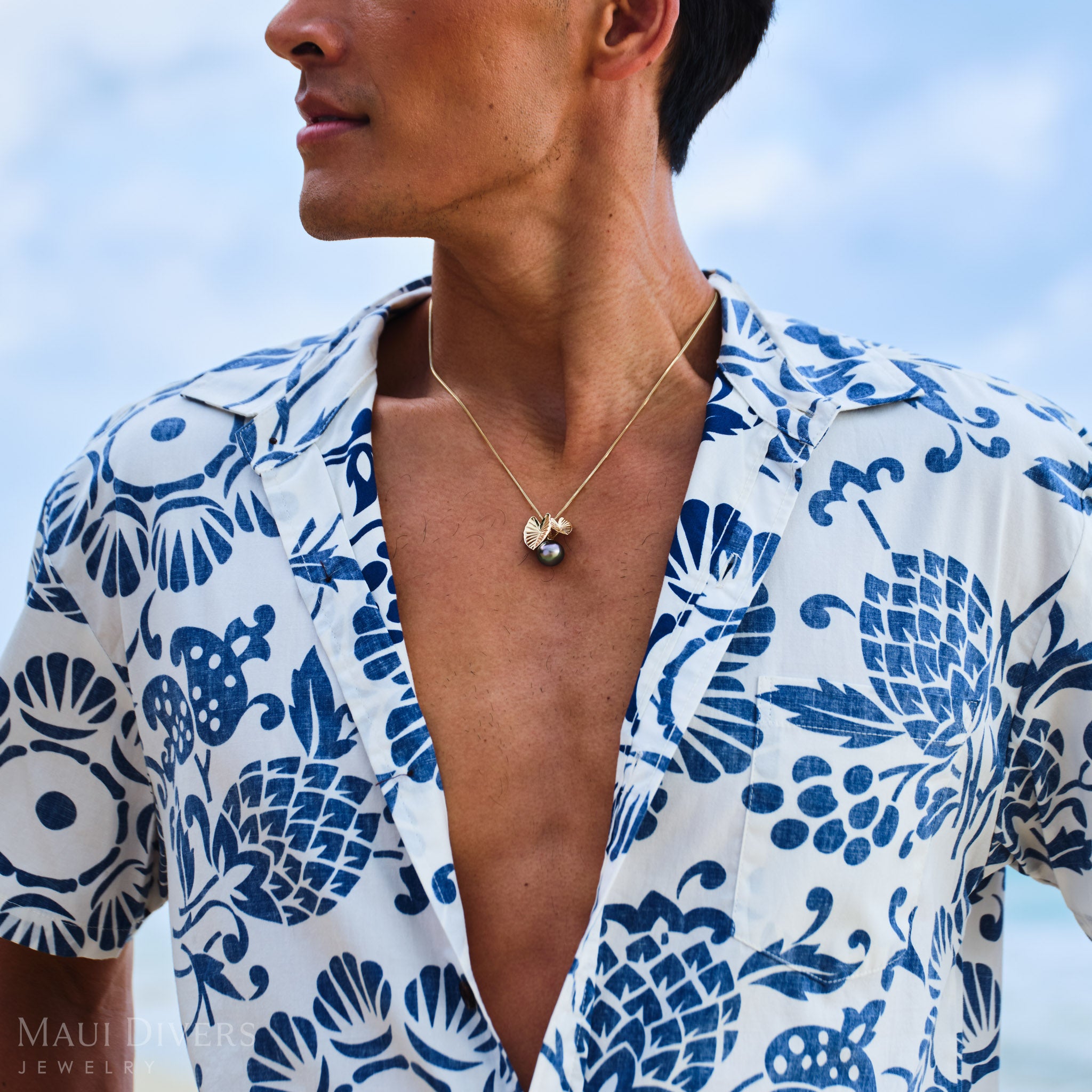 Man wearing a blue and white patterned shirt with a Kalo Tahitian black pearl necklace, outdoors.