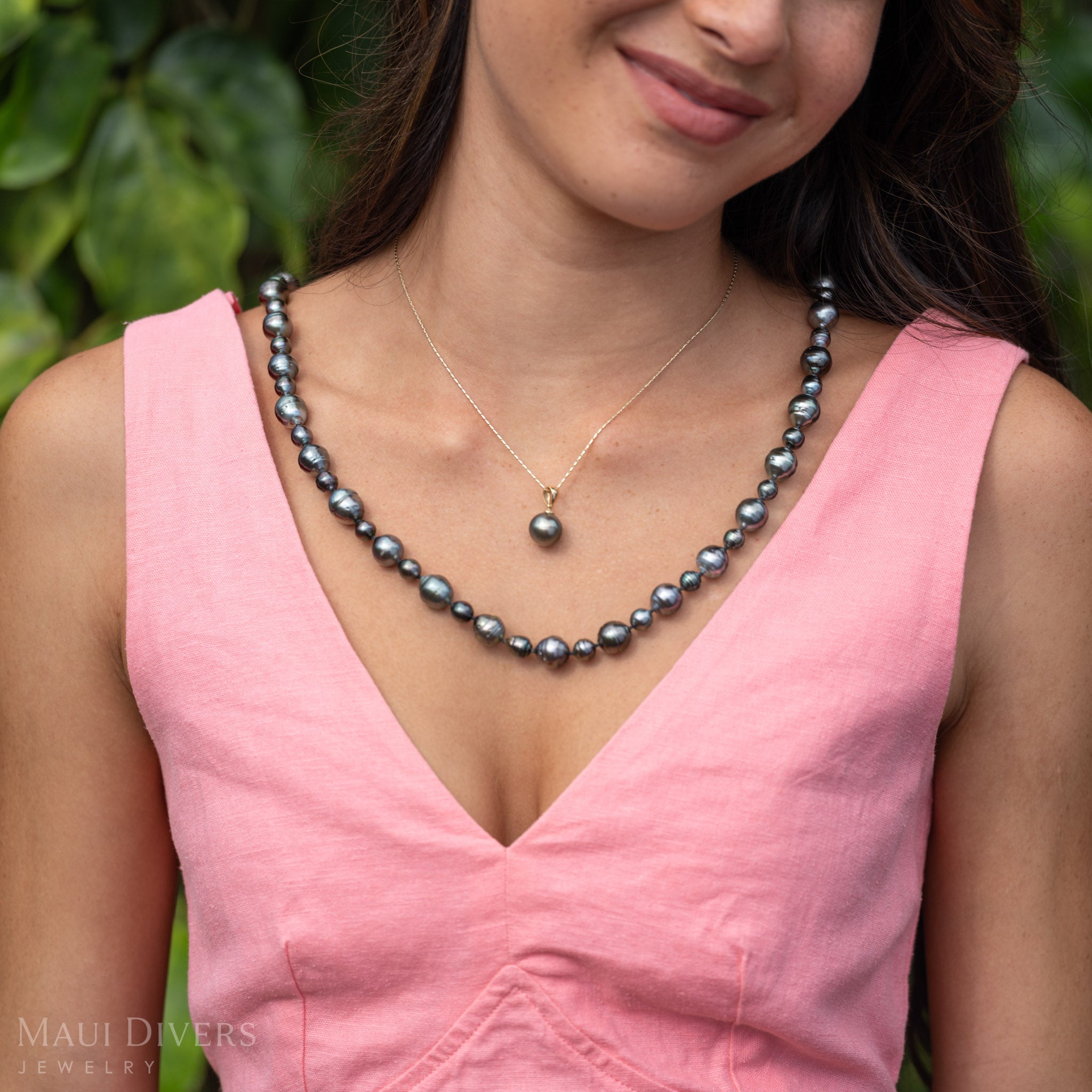 Woman wearing a Natural Tahitian Black Pearl Strand Necklace paired with a single Tahitian Black Pearl Necklace with a pink dress and green leafy blurred background. 