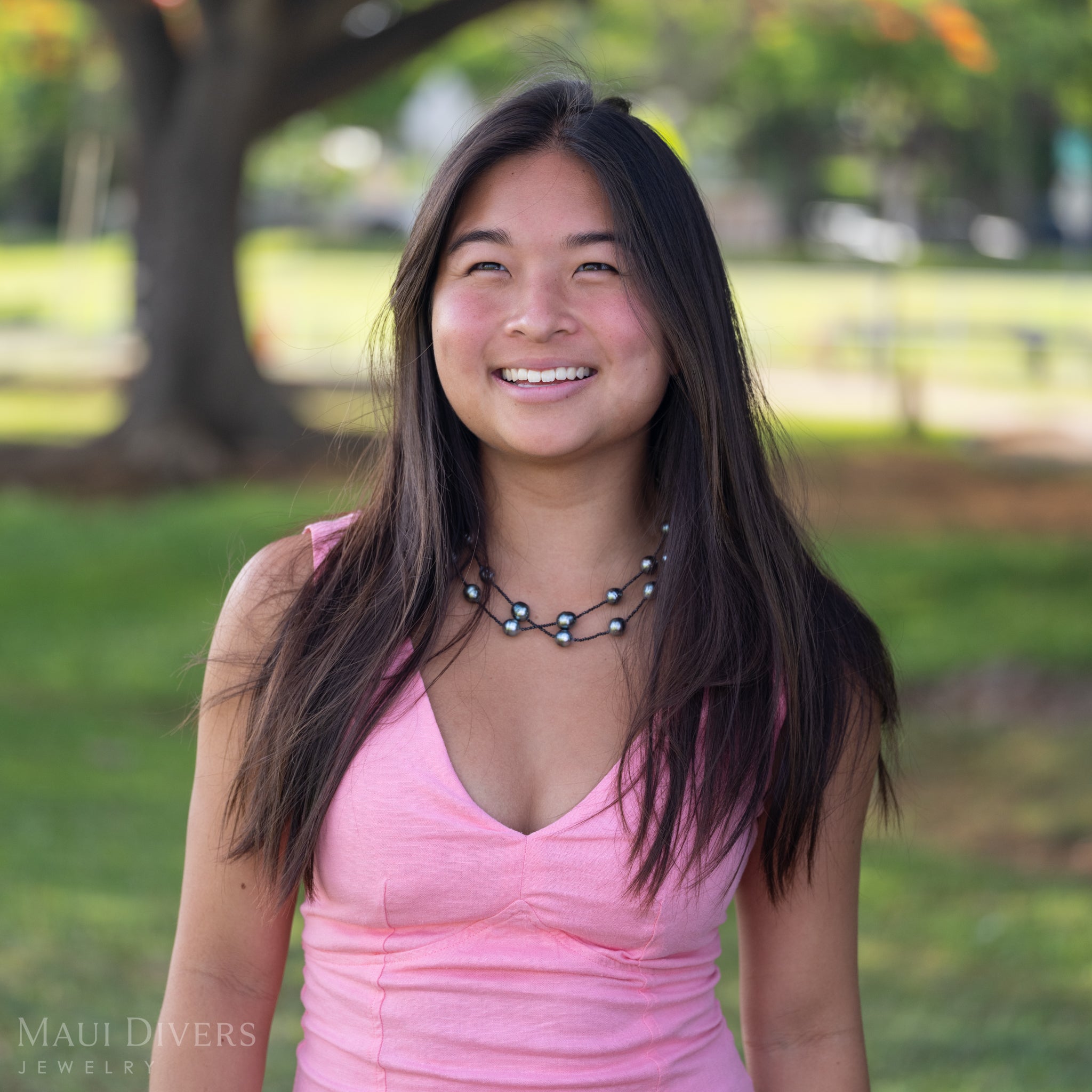 A smiling woman in a light pink top wearing a Tahitian Black Pearl and Black Spinel Layered Necklace with a 14k yellow gold clasp, against a blurred park background